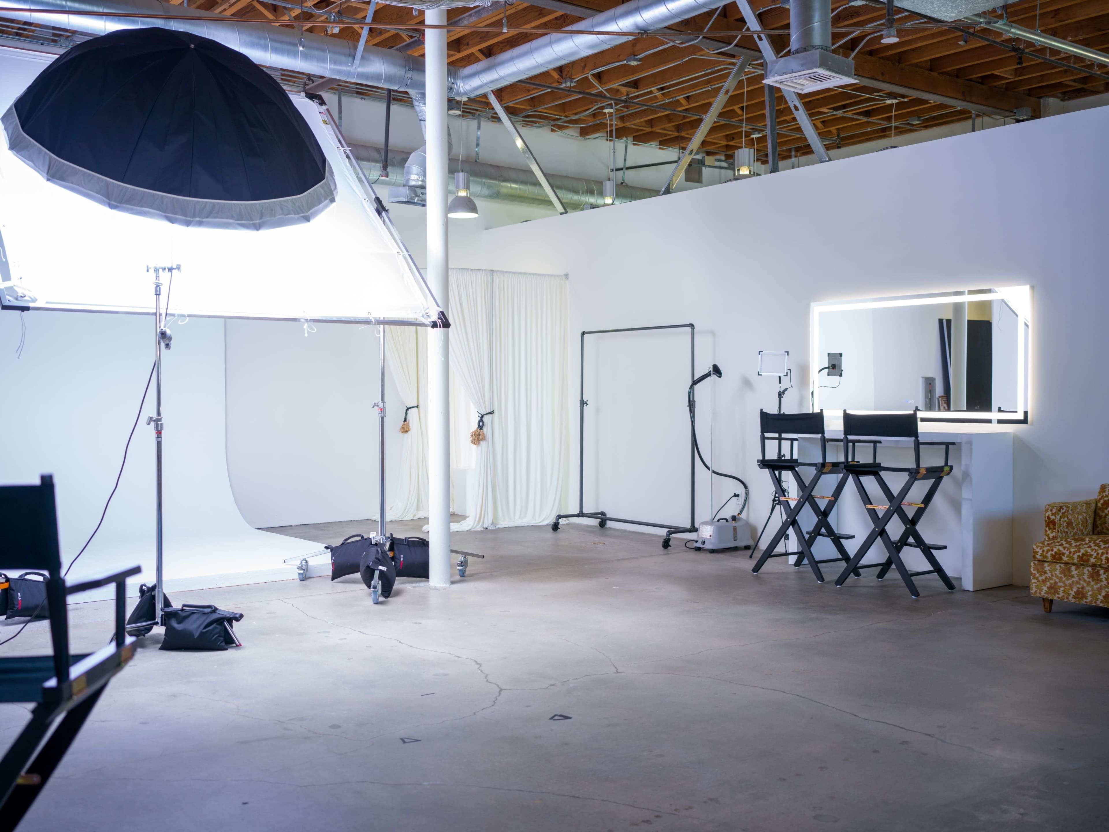 A photography studio with a large softbox light, a white backdrop, and a makeup area featuring two director's chairs and a vanity mirror.