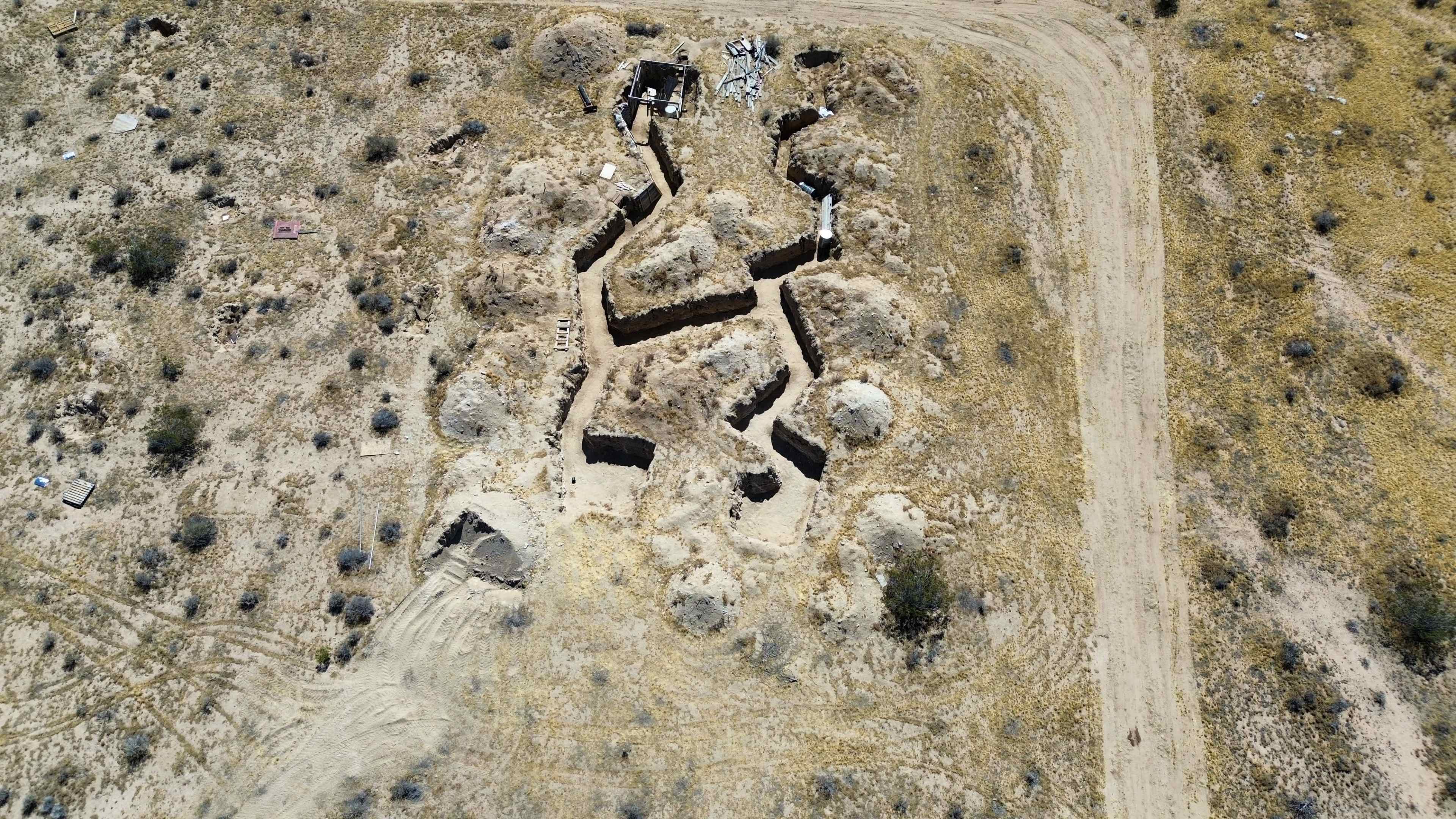 The image shows a barren landscape with a series of dug trenches and mounds, intersecting among sparse vegetation, along a dirt road.
