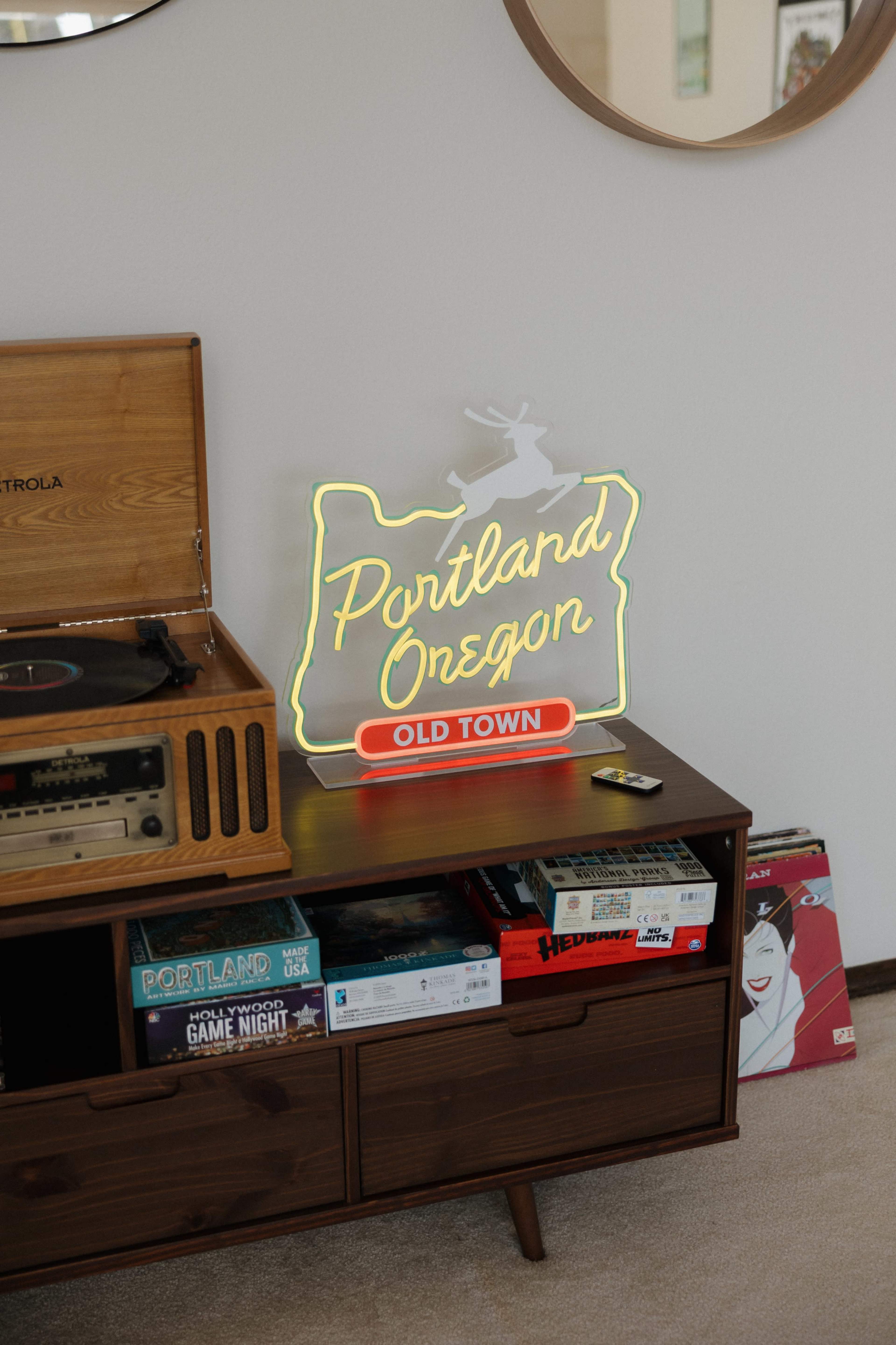 A neon sign shaped like the state of Oregon displays the words "Portland Oregon" and "OLD TOWN" on a wooden console next to a record player and various board games.