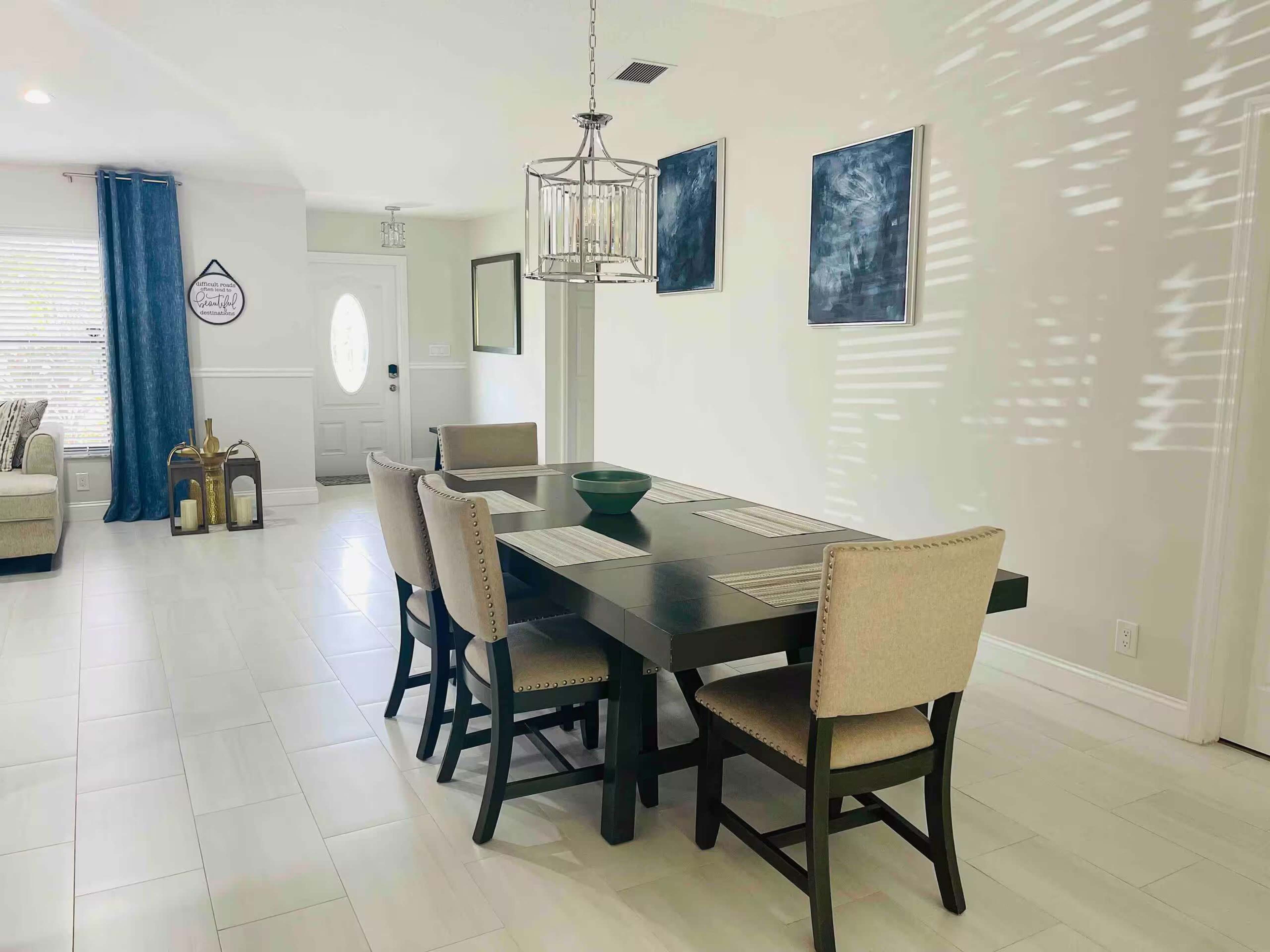 A dining area features a rectangular table with beige upholstered chairs, a chandelier above, and two abstract paintings on the walls amid a bright, minimalist space.