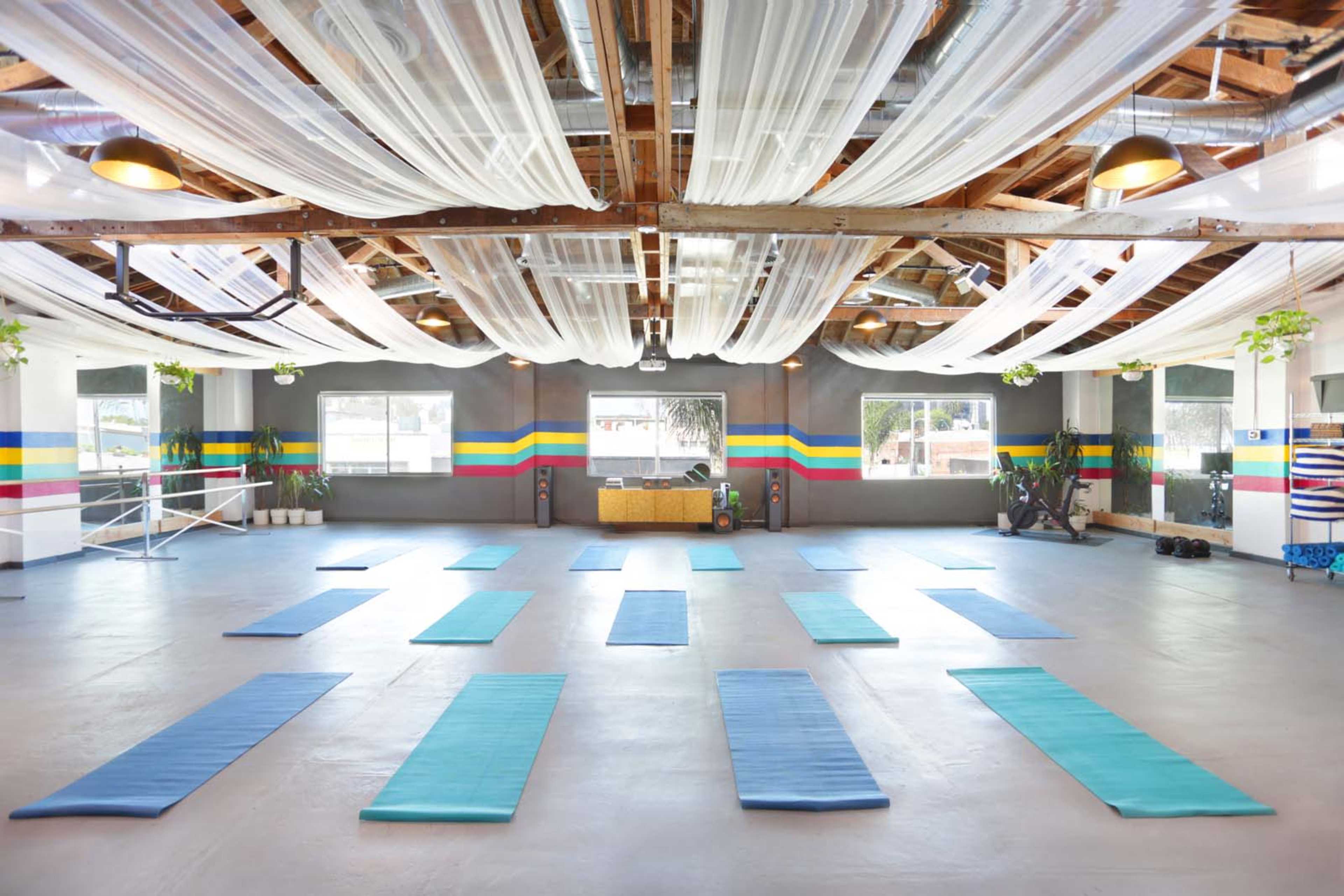 The image shows a spacious yoga studio with blue mats arranged neatly in rows on a gray floor, adorned with colorful striped walls and soft lighting from above.