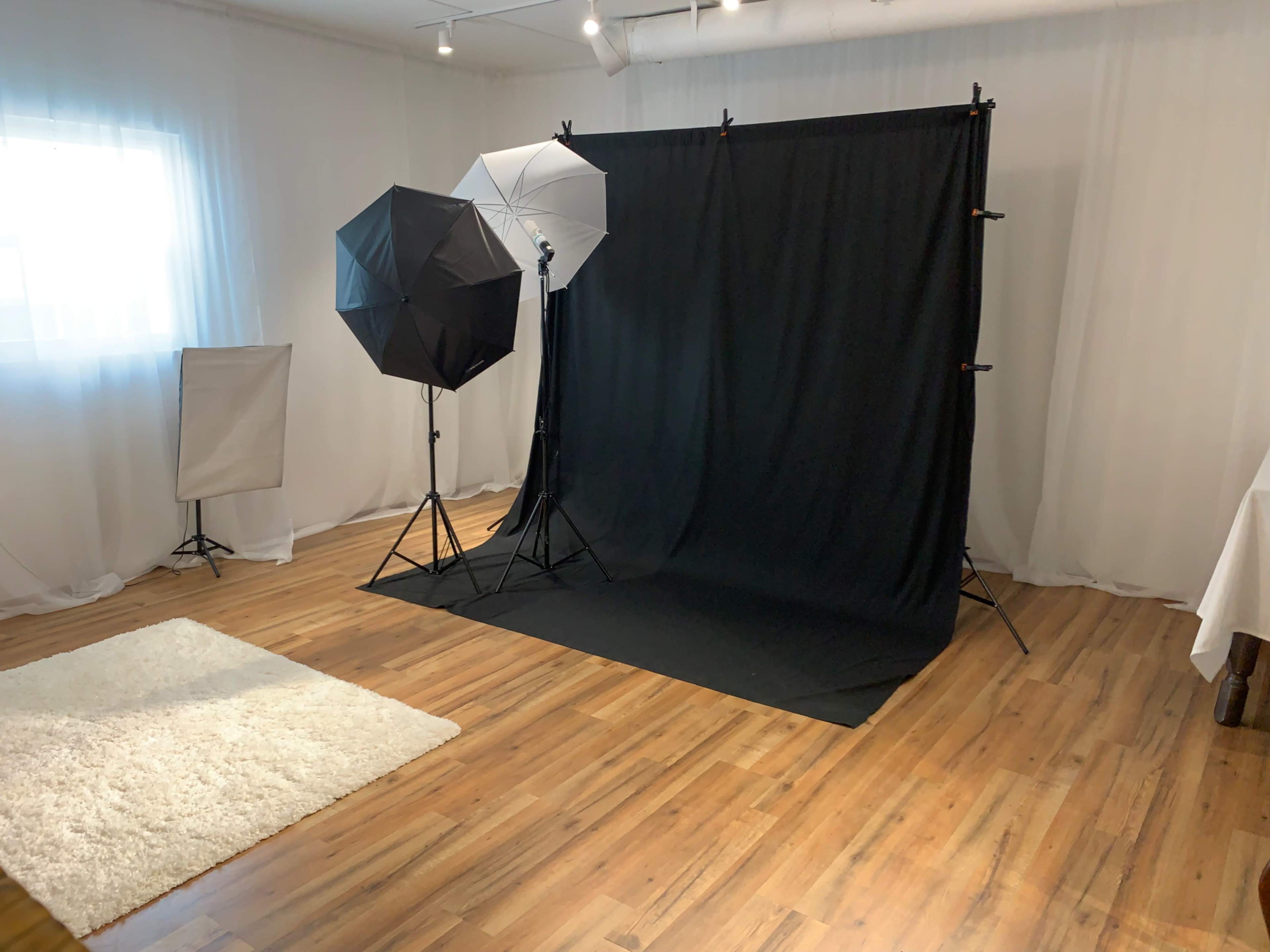 A photography studio is set up with a black backdrop, two softbox lights, and a white rug on a wooden floor.