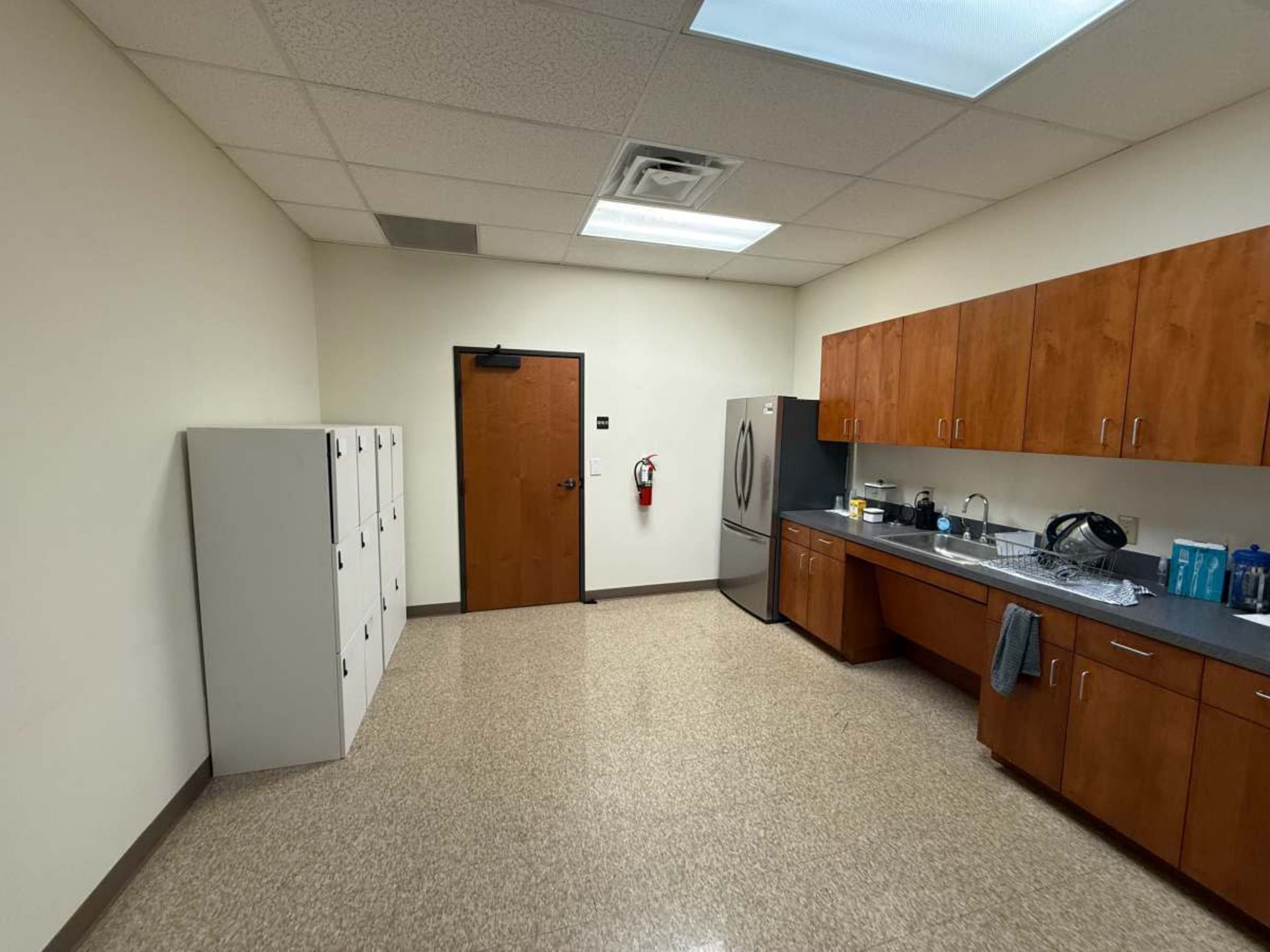 The image shows a break room with wooden cabinets, a refrigerator, and a row of lockers along one wall.