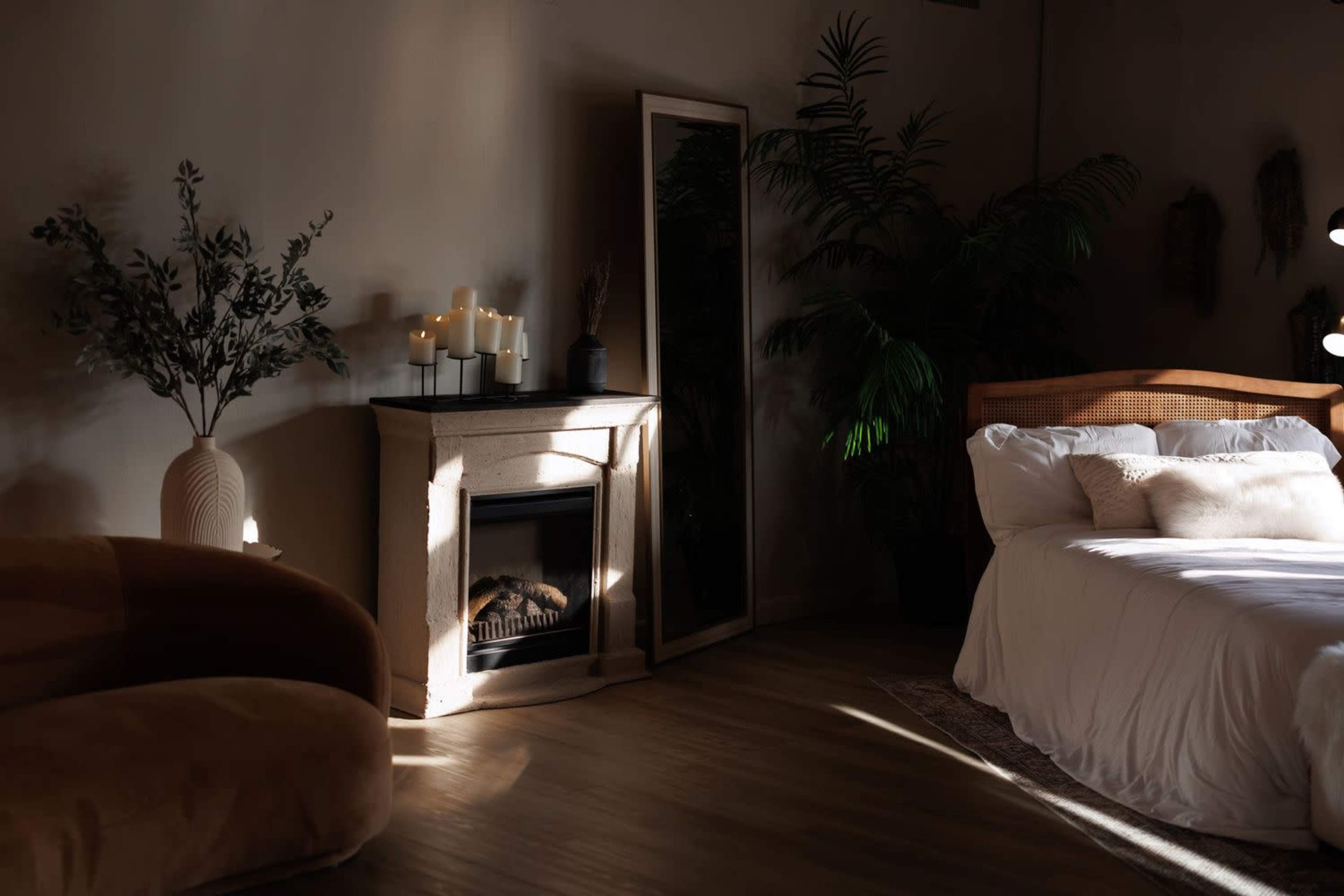 A cozy bedroom features a bed with white linens, a fireplace with candles, a large mirror, and a plant in the corner.
