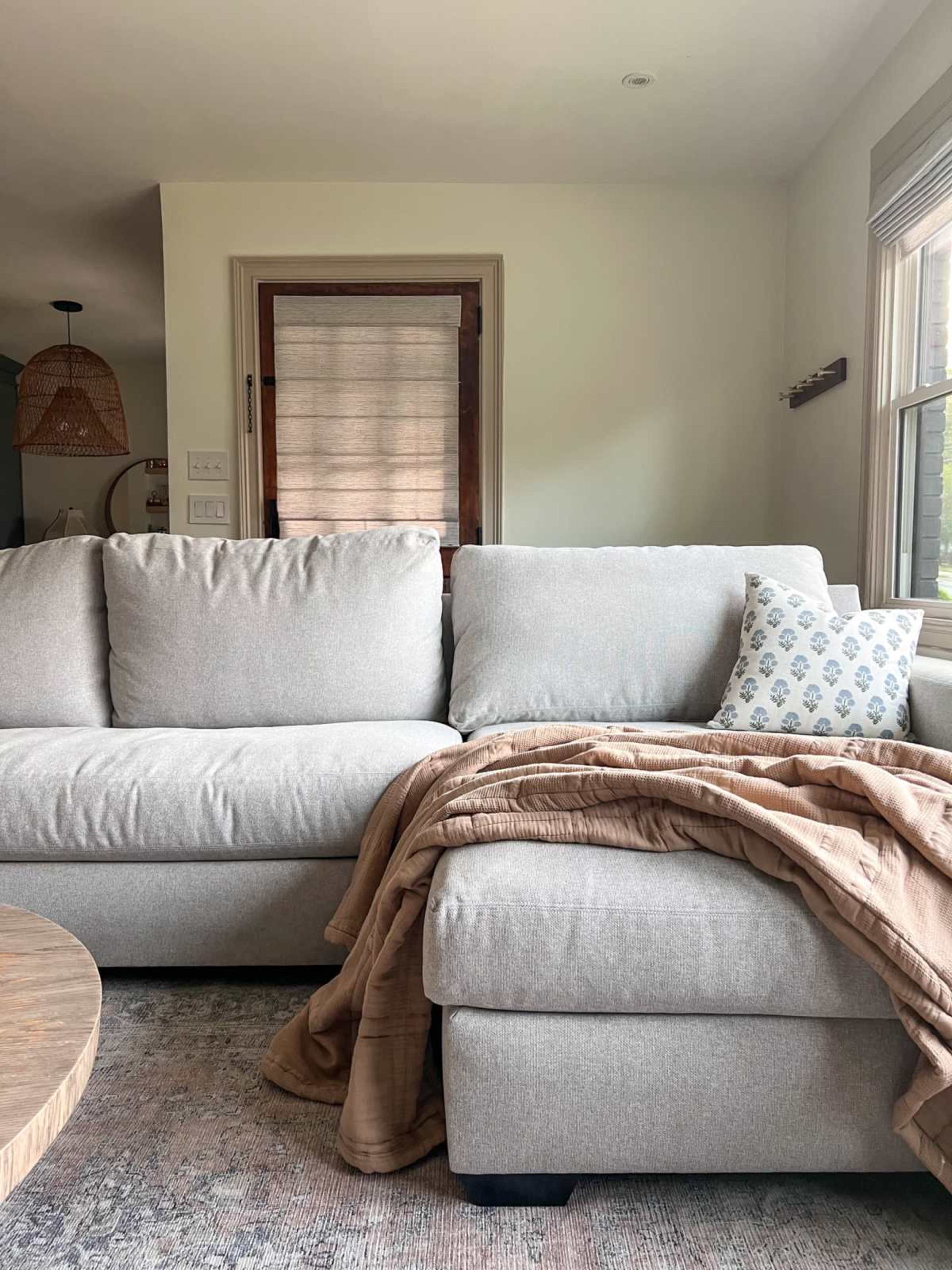 A light gray sectional couch with a patterned throw pillow and a brown blanket is positioned in a cozy living room near a wooden coffee table and windows.