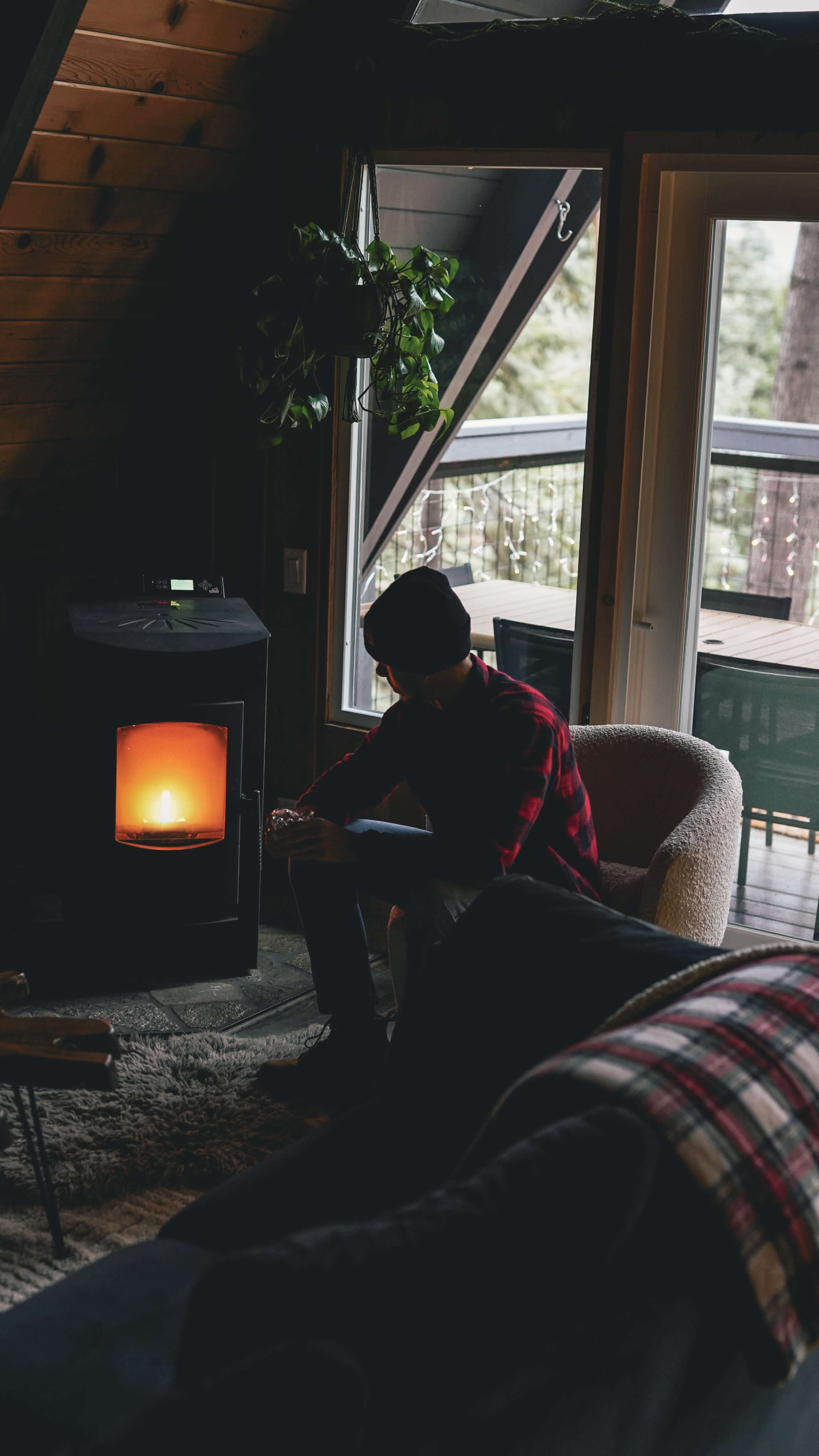 A person in a red plaid shirt sits in a chair near a glowing stove in a cozy room with large windows.