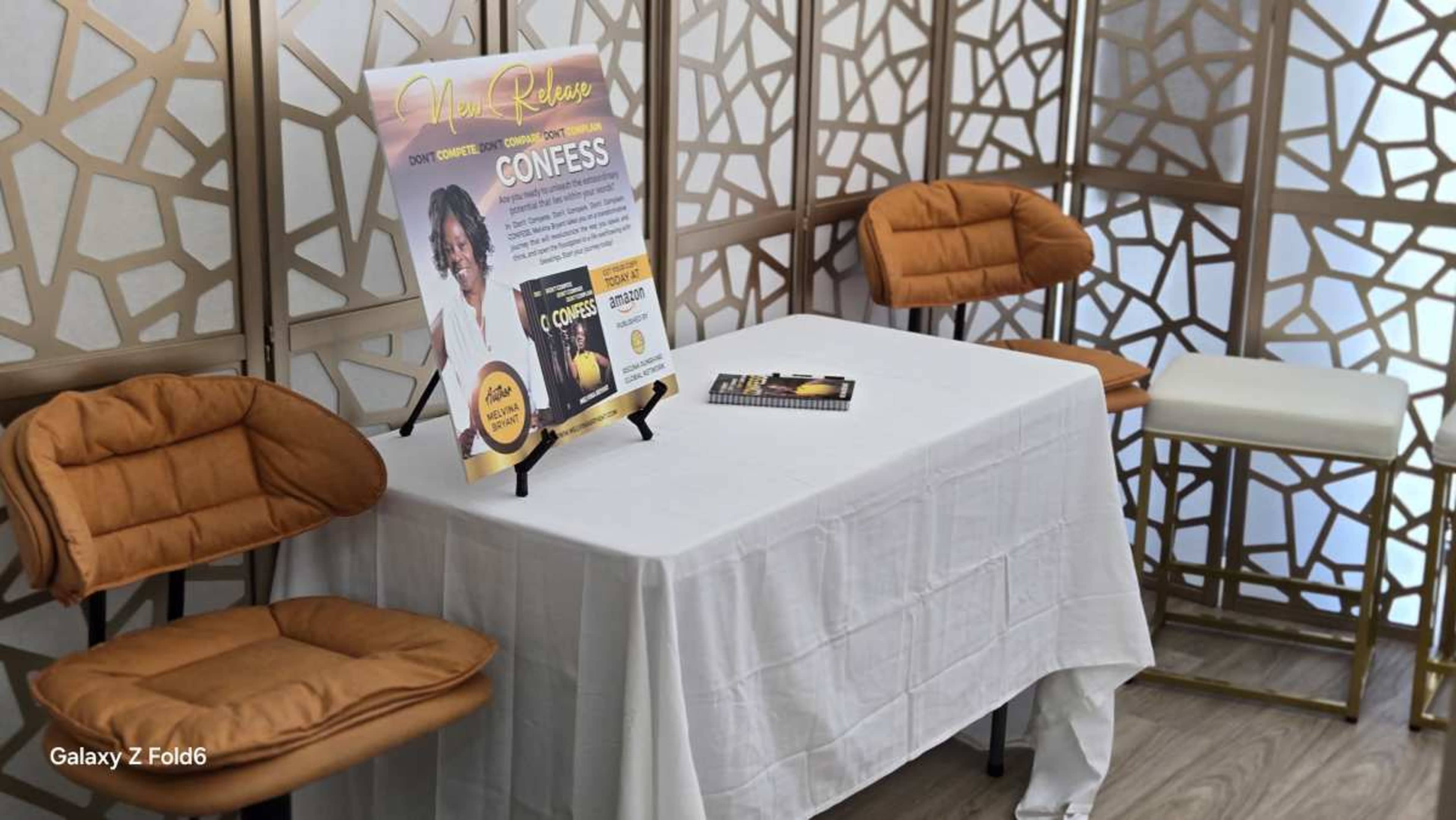 A small table covered with a white tablecloth displays a promotional sign for an event titled "Confess," accompanied by two orange chairs and a white chair against a decorative background.
