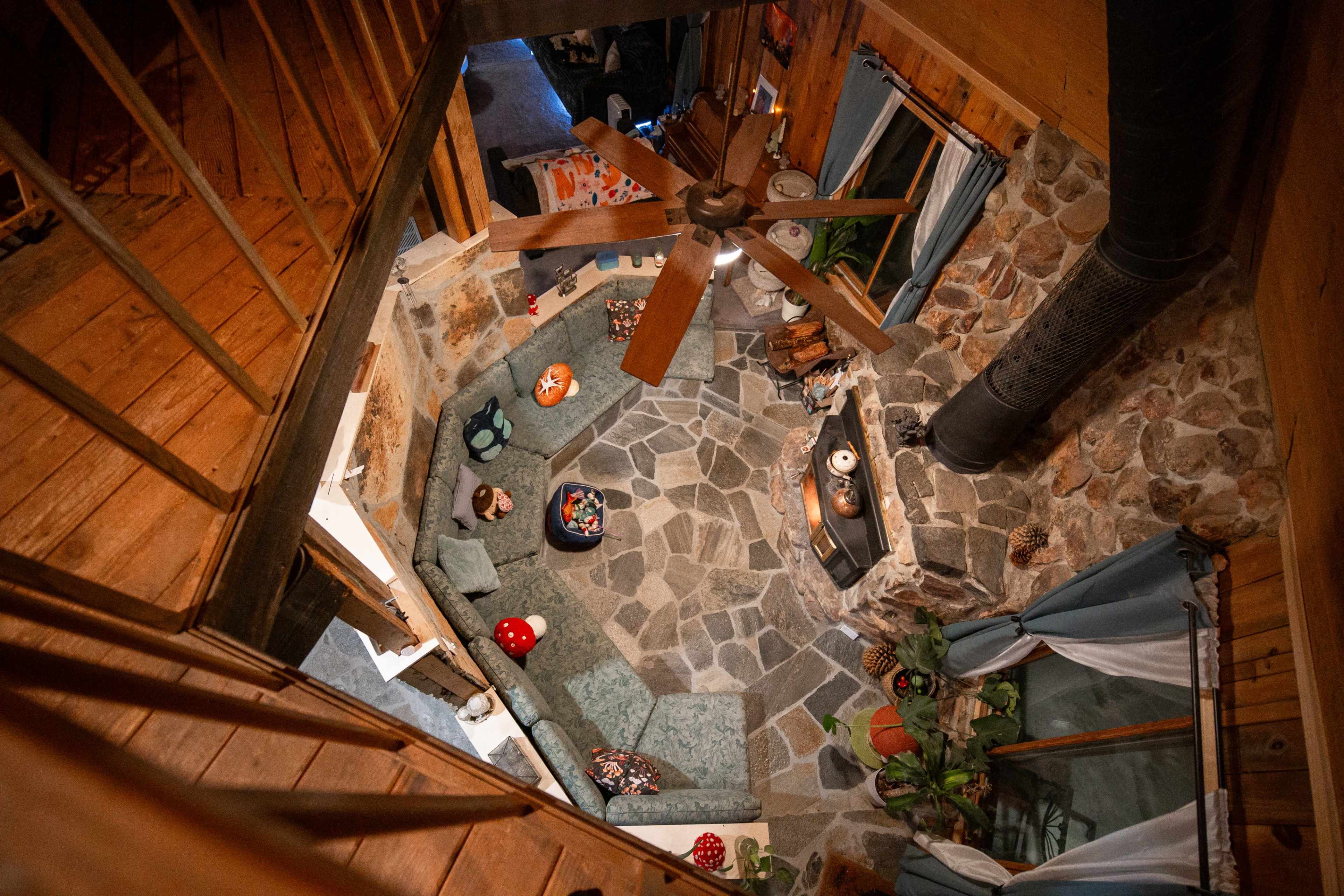 The image shows an overhead view of a cozy living room featuring a stone floor, a fireplace, and wooden beams, with a fan hanging from the ceiling.