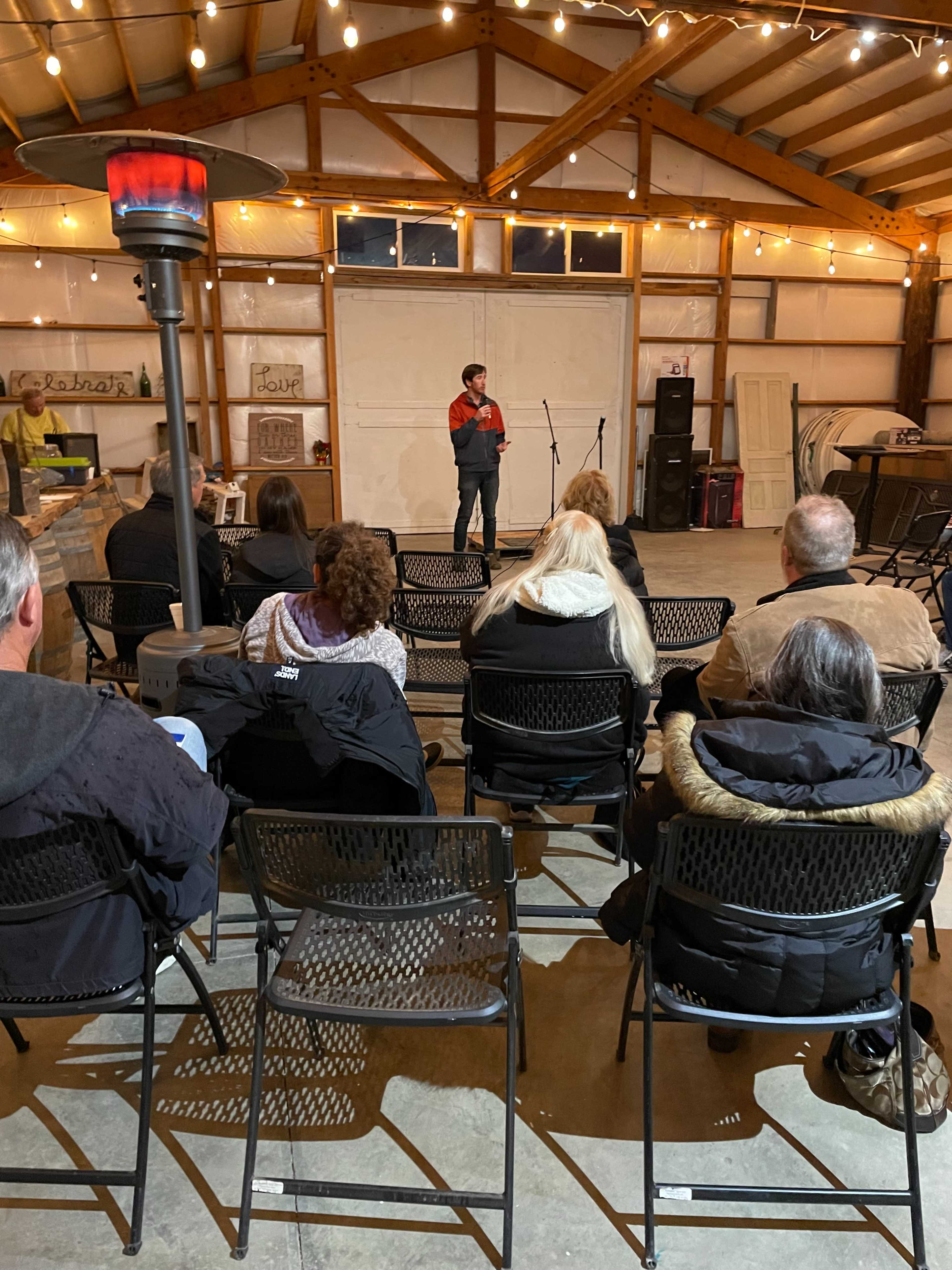 A speaker stands on a small stage in a barn-like venue while an audience sits in rows of chairs, illuminated by string lights above.