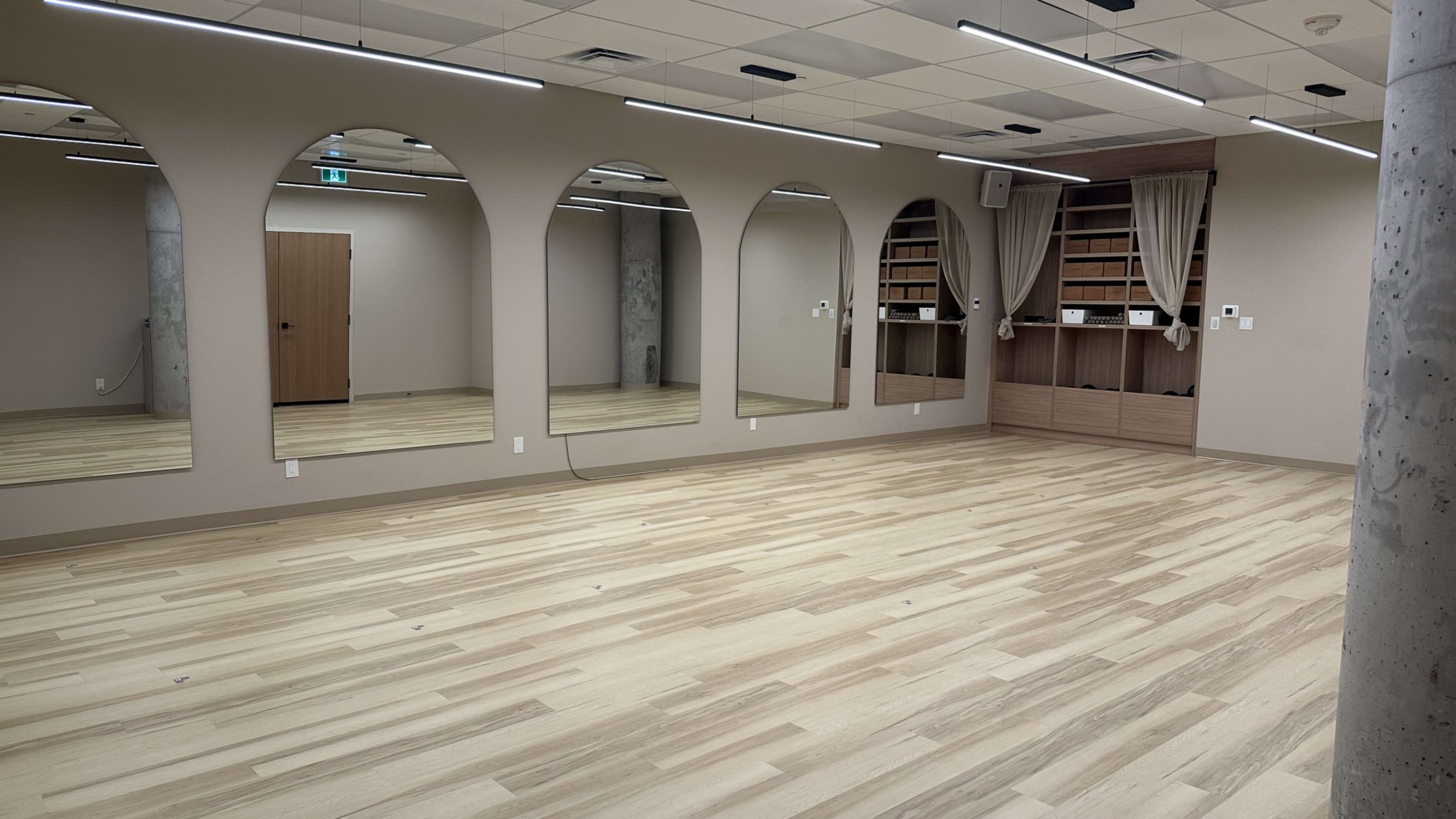 The image shows a spacious indoor room with light wood flooring, three large arched mirrors on one wall, and shelving along another wall.