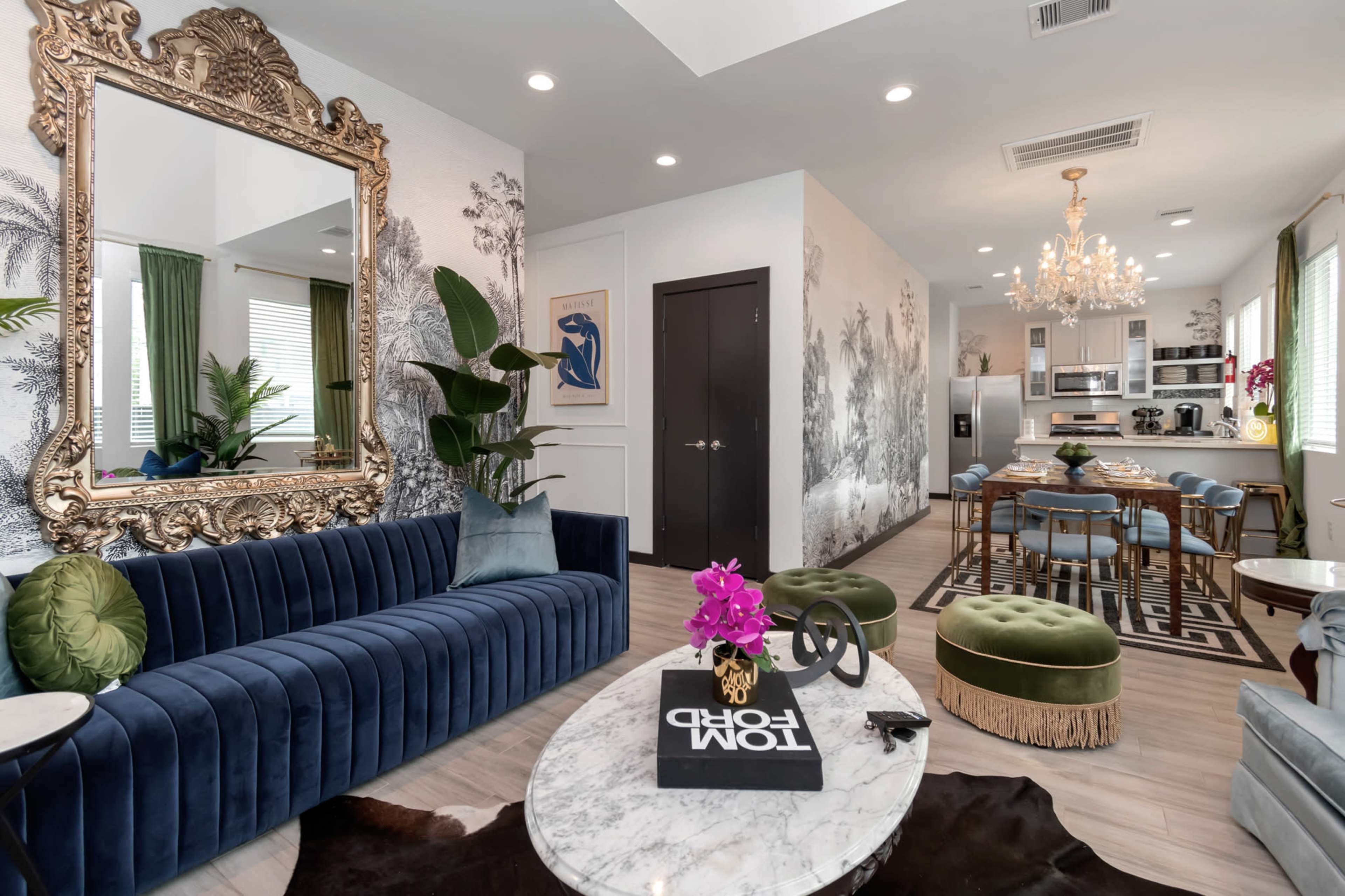 A modern living space featuring a blue velvet sofa, a large ornate mirror, a marble coffee table, and a dining area with a chandelier.