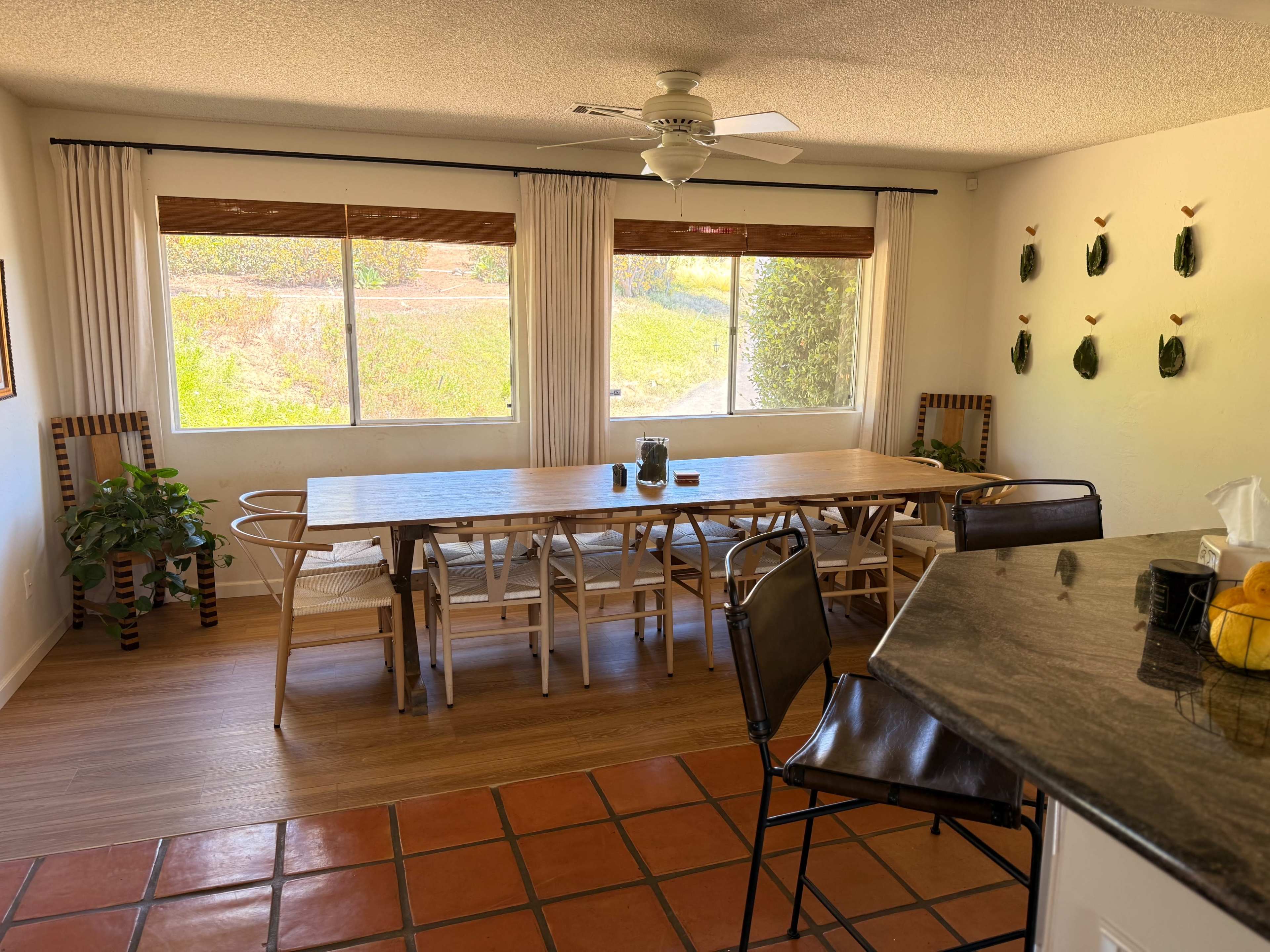 The image shows a spacious dining area with a large wooden table, light-colored walls, and large windows providing natural light, complemented by a ceiling fan and decorative wall hangings.