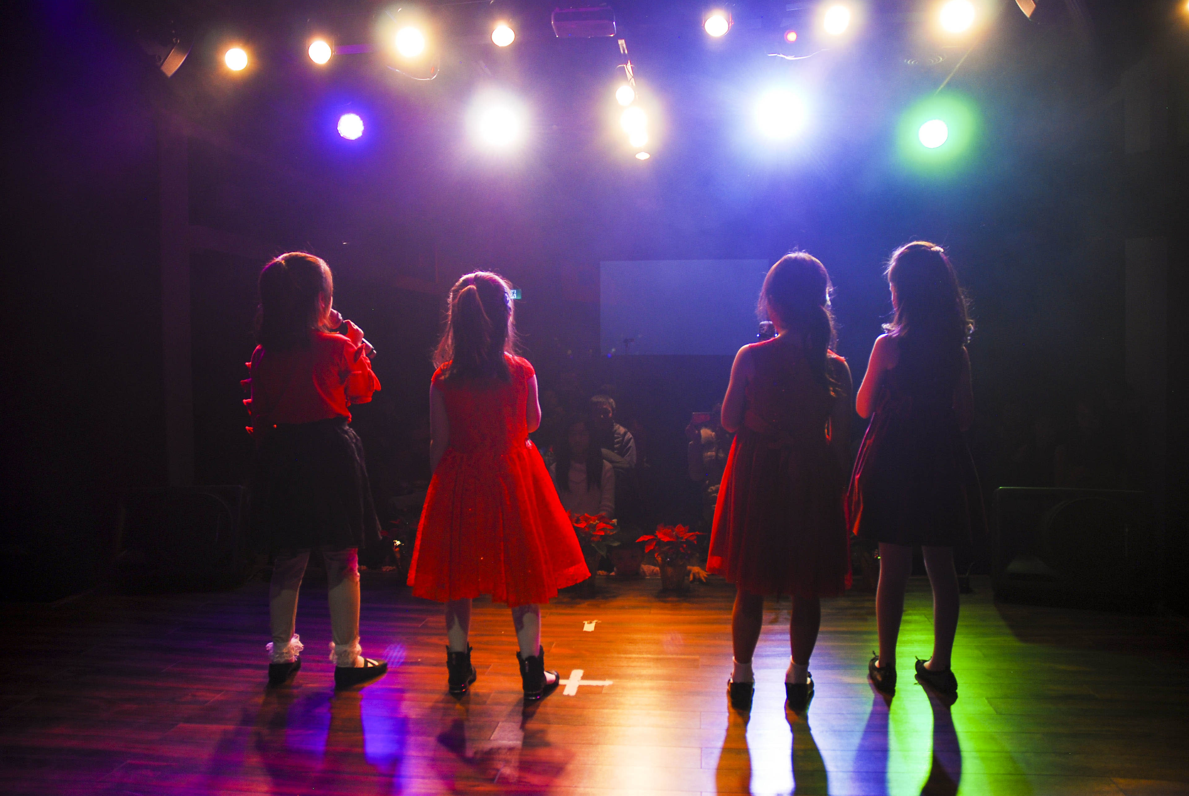 A group of four young girls, dressed in colorful dresses, stands on a stage with bright lights shining above them.