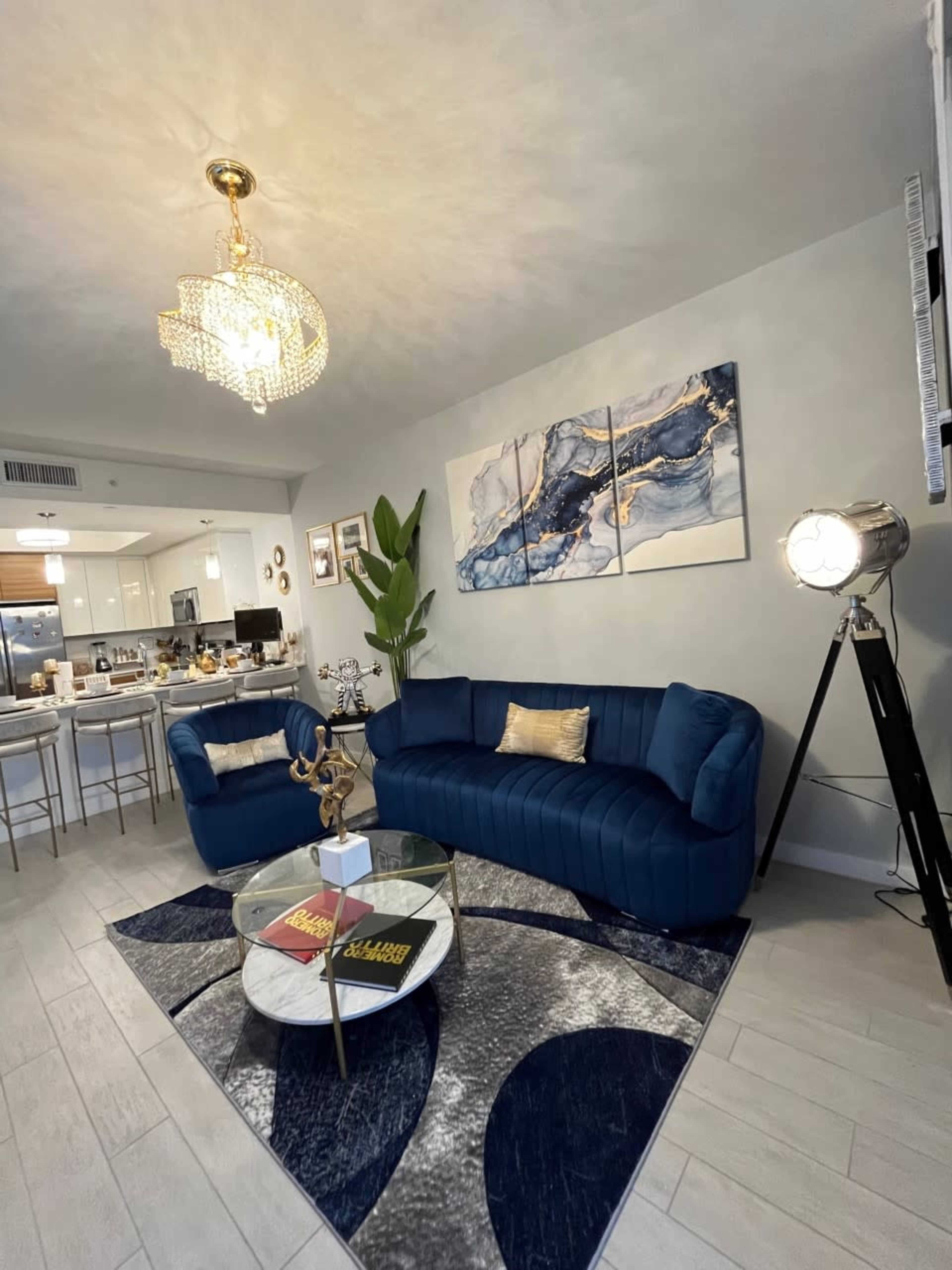 The image shows a modern living room featuring a blue couch, a circular coffee table with books, and a decorative light fixture, with a kitchen area visible in the background.