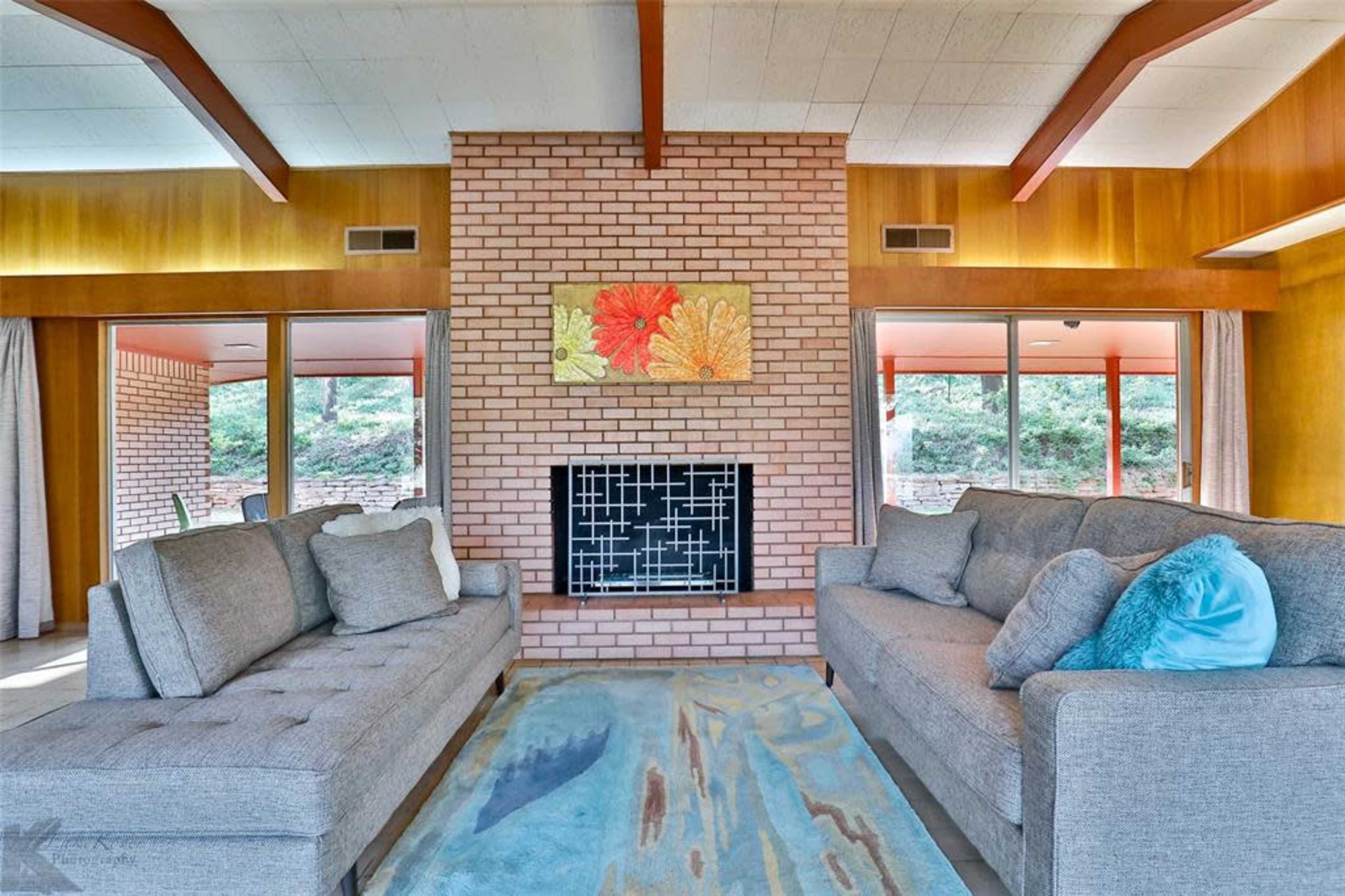 The room features two gray sofas set opposite a brick fireplace, with large windows that provide a view of the outdoors.