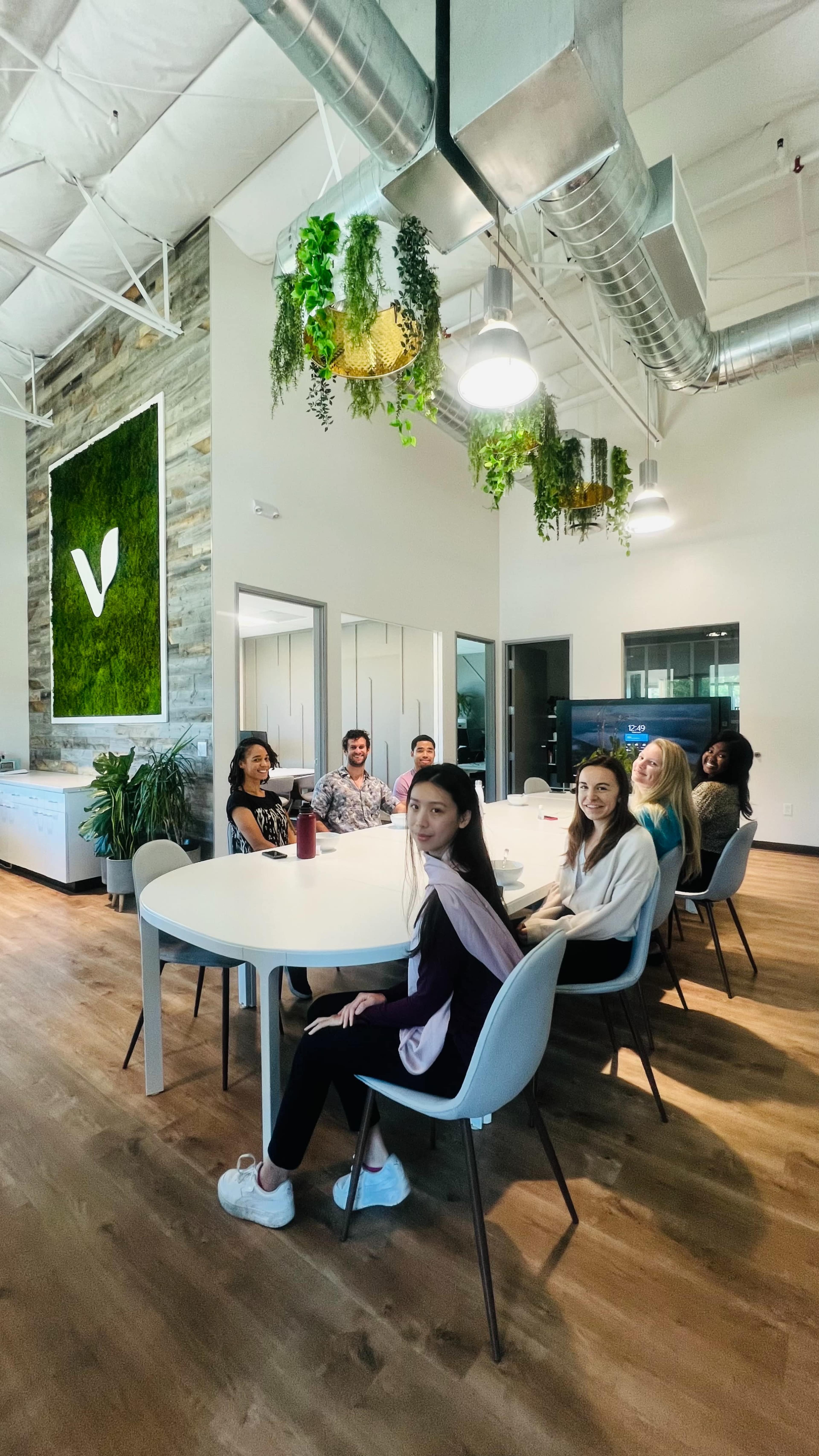 A group of people is sitting around a large, round table in a well-lit modern office with greenery on the walls and various plants around the room.