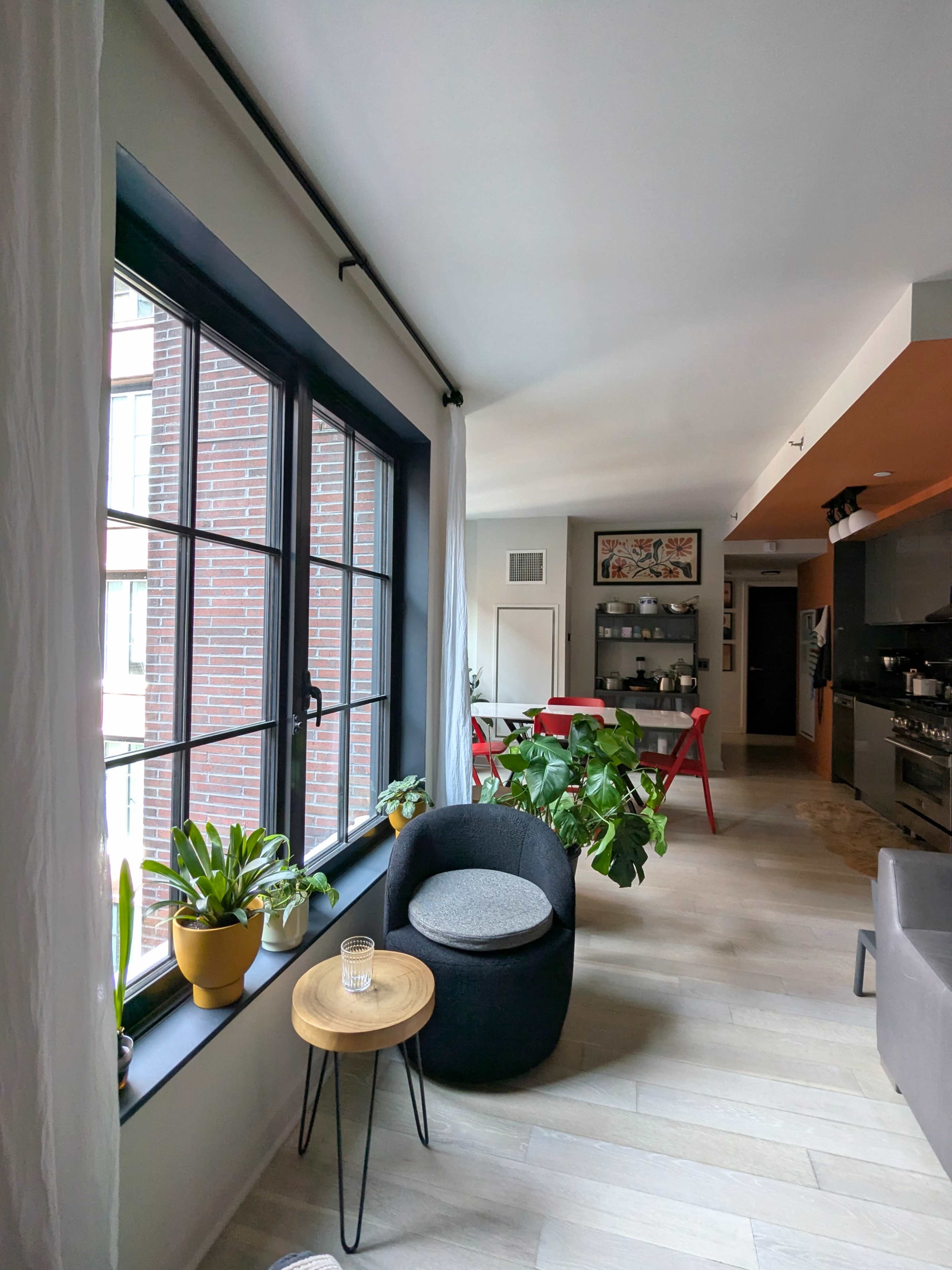 A modern living space features a cozy chair beside a small table, with large windows allowing natural light and greenery visible in the corner.