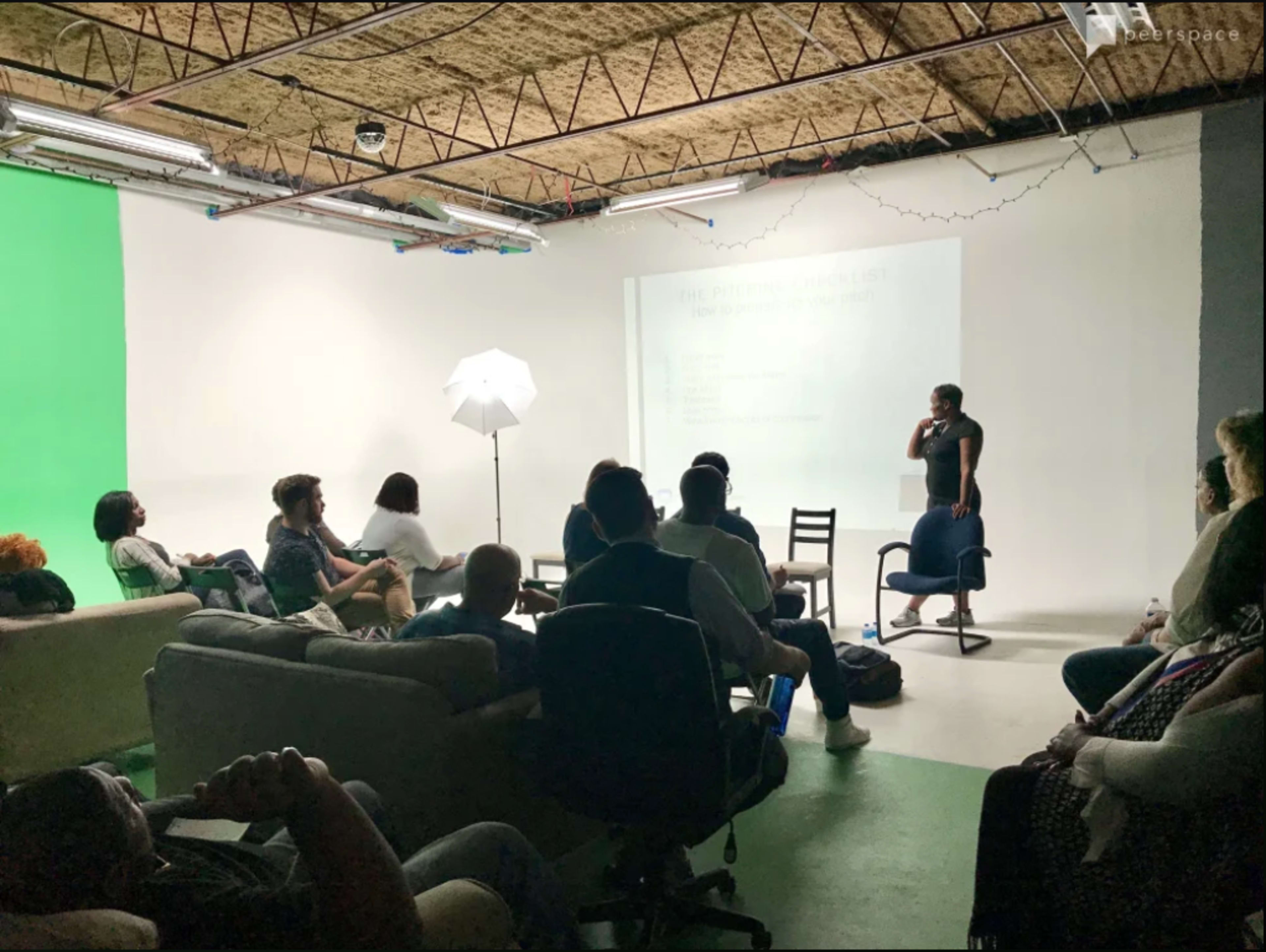 A speaker presents information in a studio setting to an audience seated on various furniture.