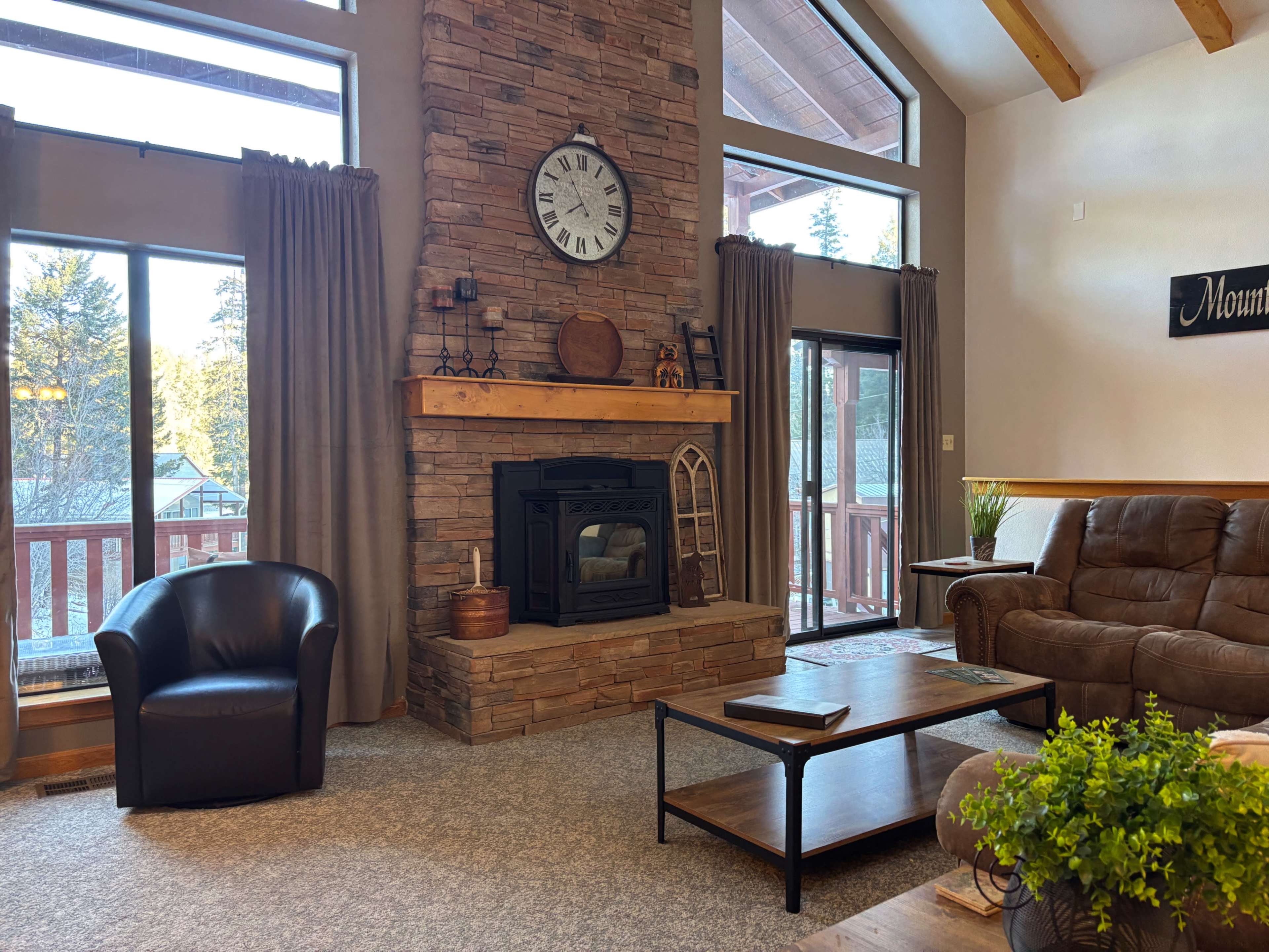The image shows a cozy living room featuring a stone fireplace, large windows with curtains, a leather chair, and a wooden coffee table.