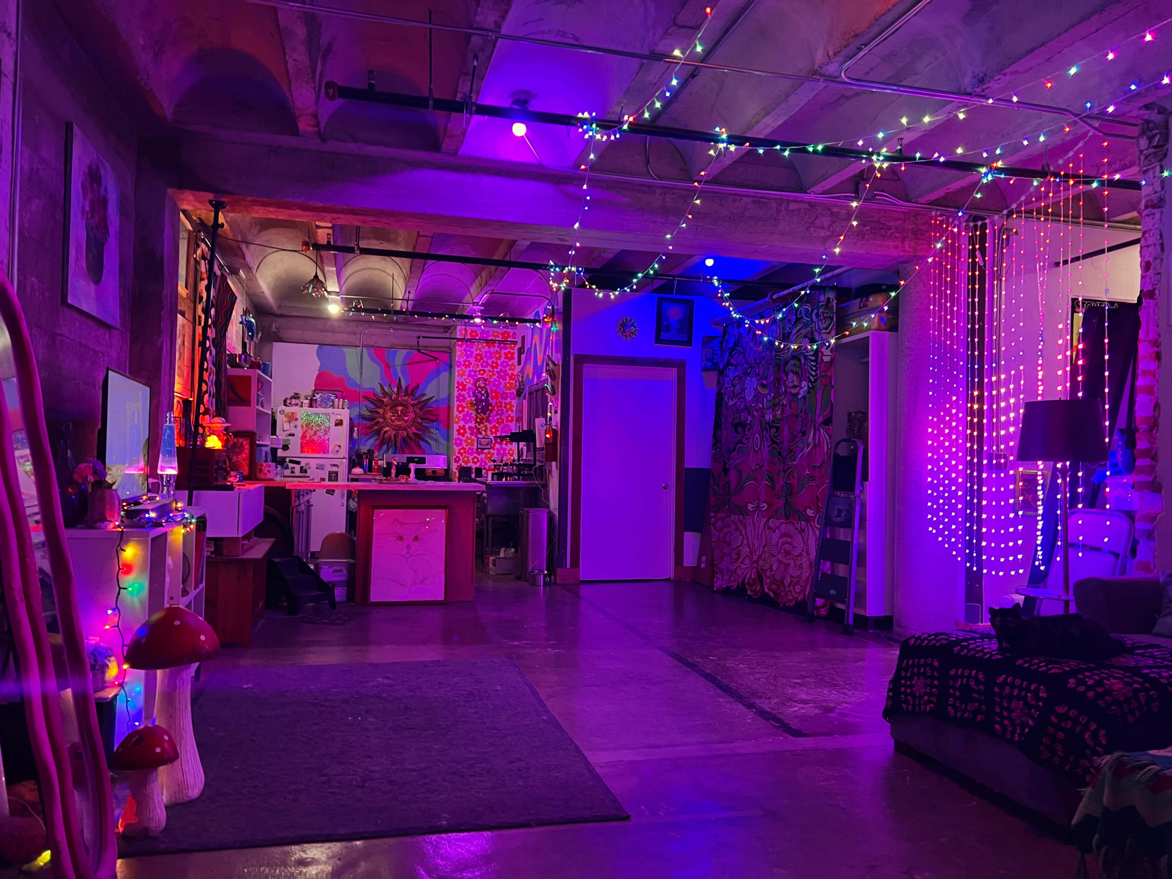 The image shows a brightly lit room with colorful string lights hanging from the ceiling, featuring a mix of artistic decorations and furniture.