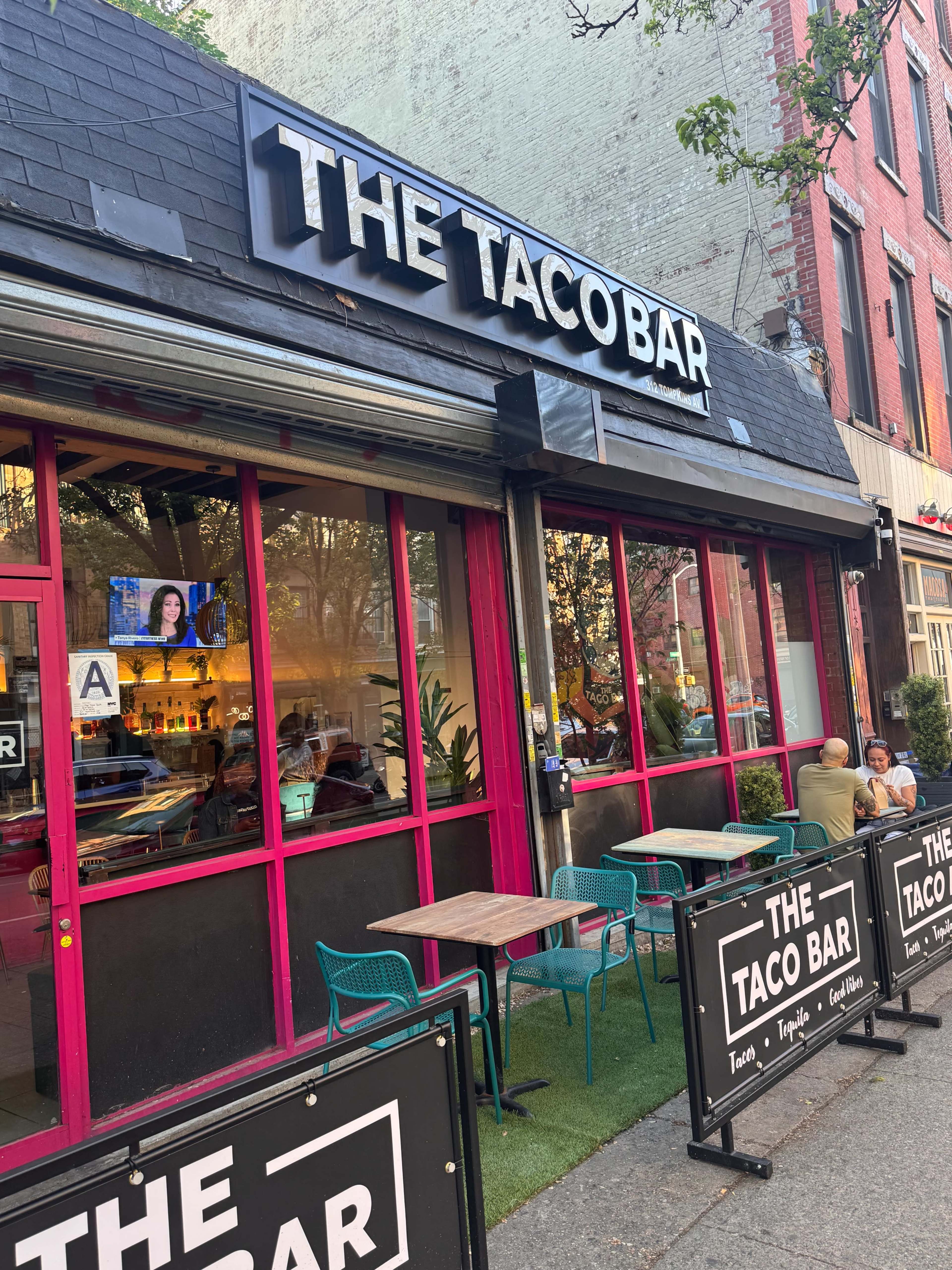 The image shows a restaurant called "The Taco Bar" with a modern exterior that features large windows, outdoor seating, and signage above the entrance.