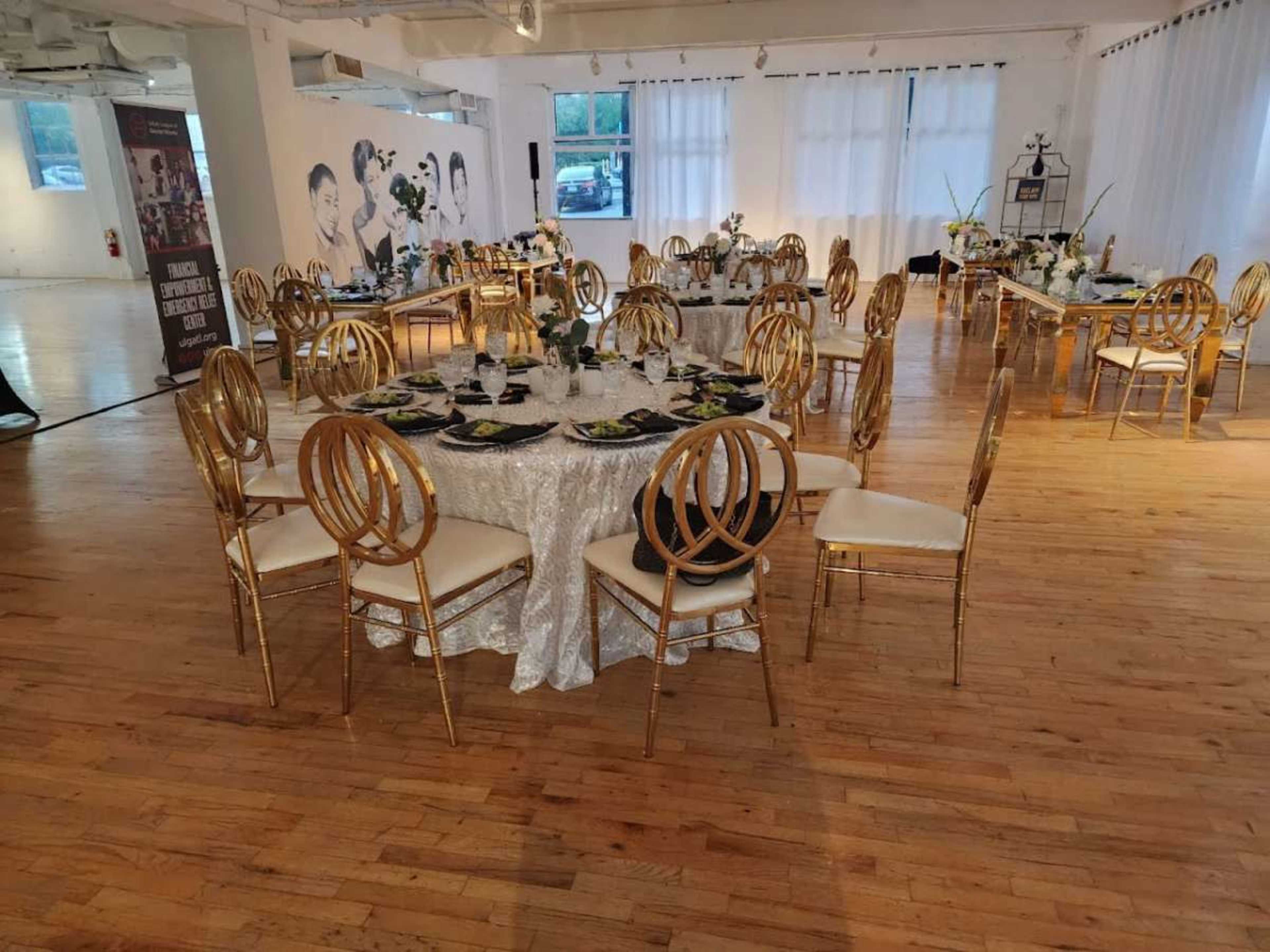 The image shows a dining hall set up for an event, featuring multiple tables with gold chairs, white tablecloths, and floral arrangements.