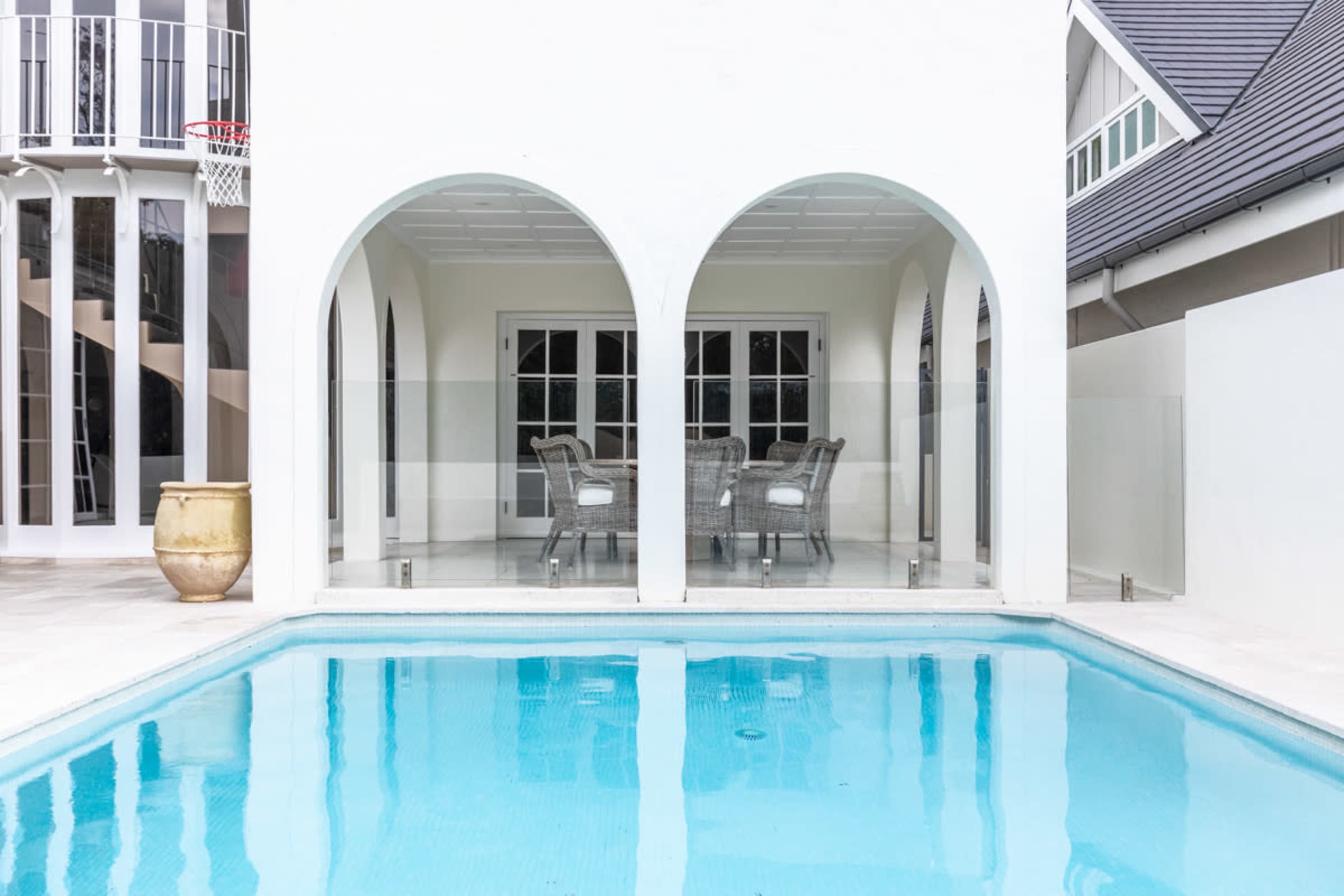 The image shows a swimming pool bordered by a white building featuring arched openings that lead to a glass-enclosed dining area.