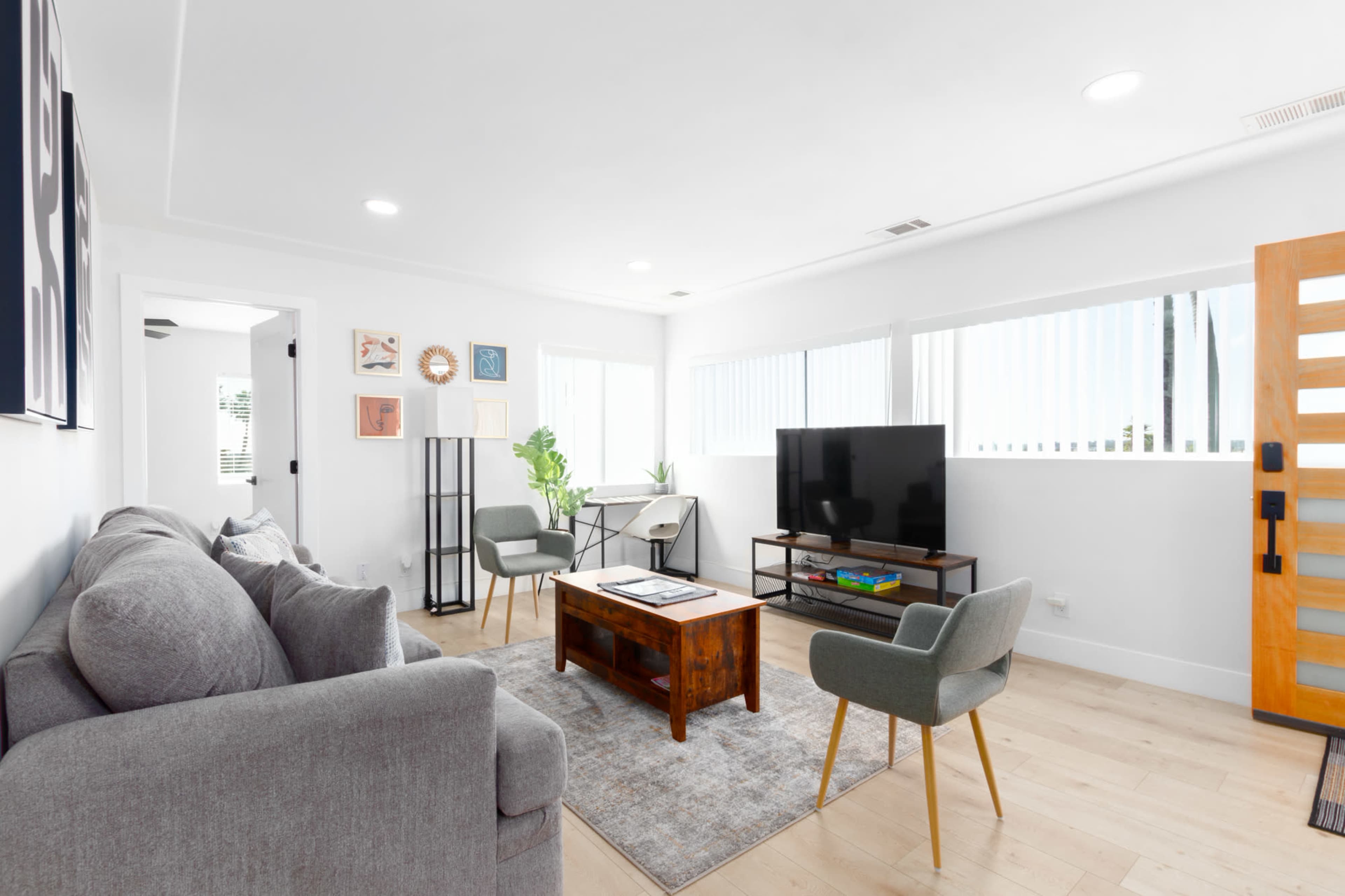 A modern living room features a gray sofa, two green chairs, a wooden coffee table, a TV stand with a television, and large windows with blinds.