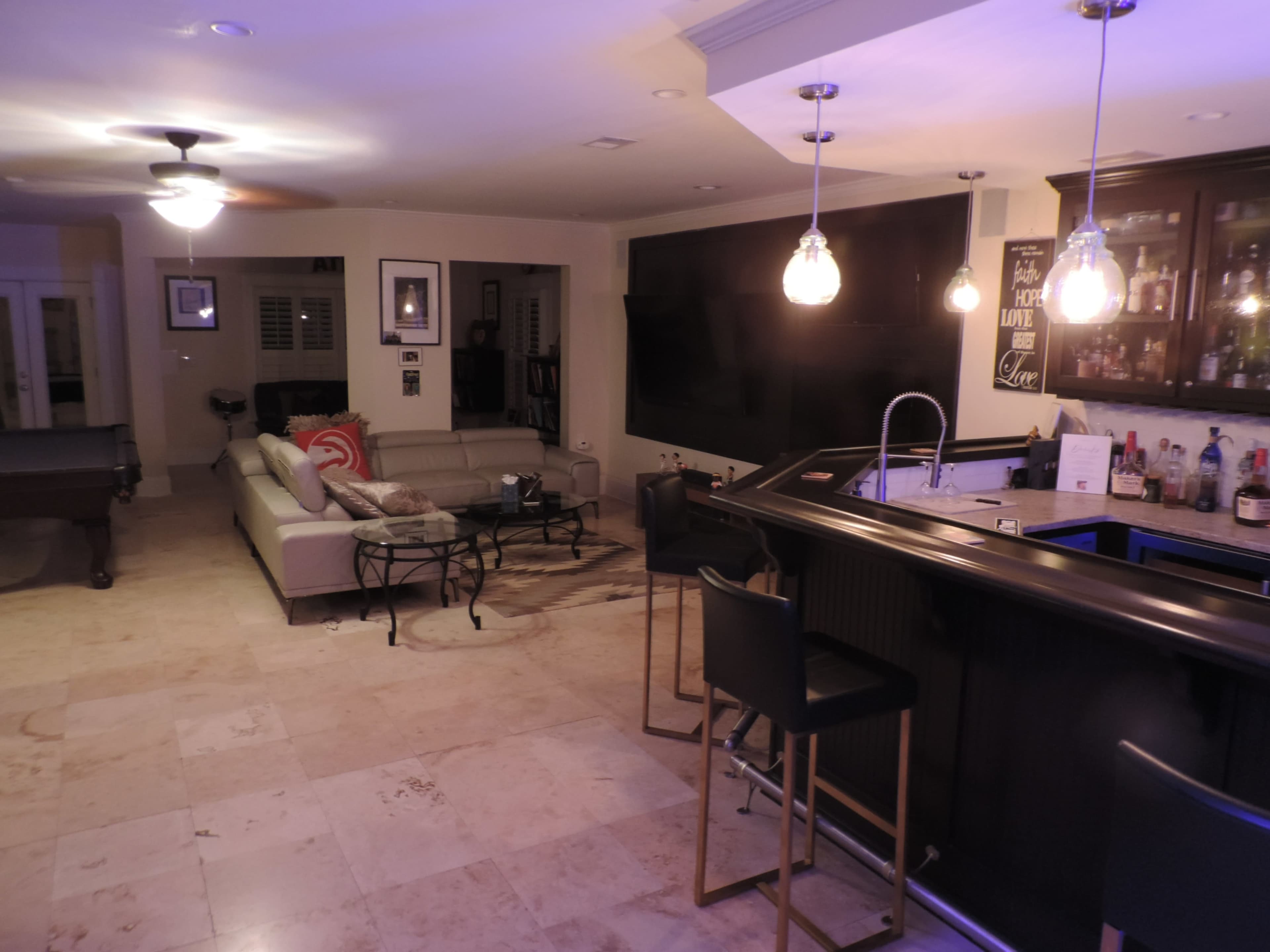 The image shows a spacious living area featuring a bar with high stools, a large television on the wall, a sectional sofa, and tile flooring.