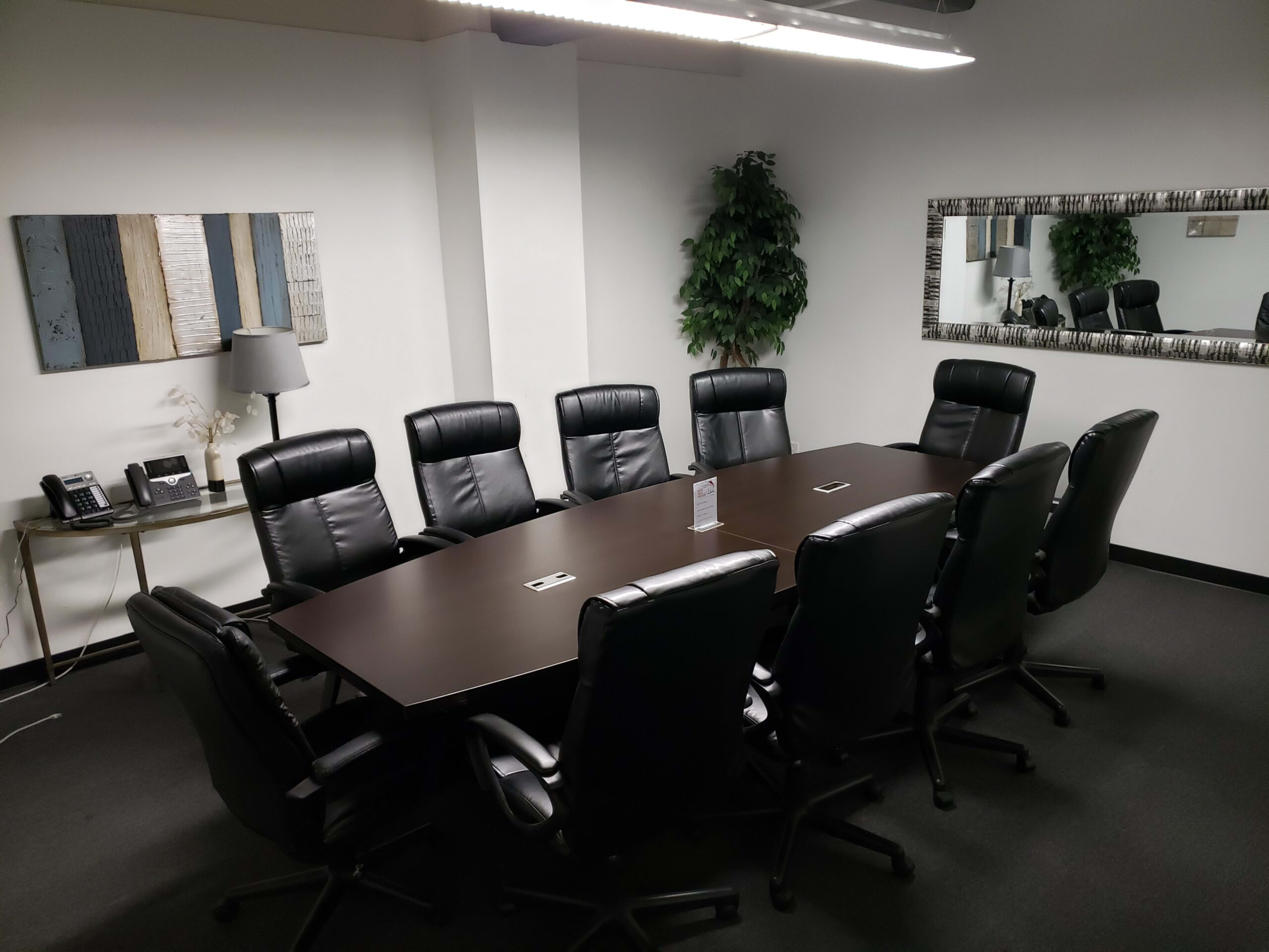 The image shows a modern conference room with a large brown table surrounded by black leather chairs, a mirror on one wall, and artwork on another.