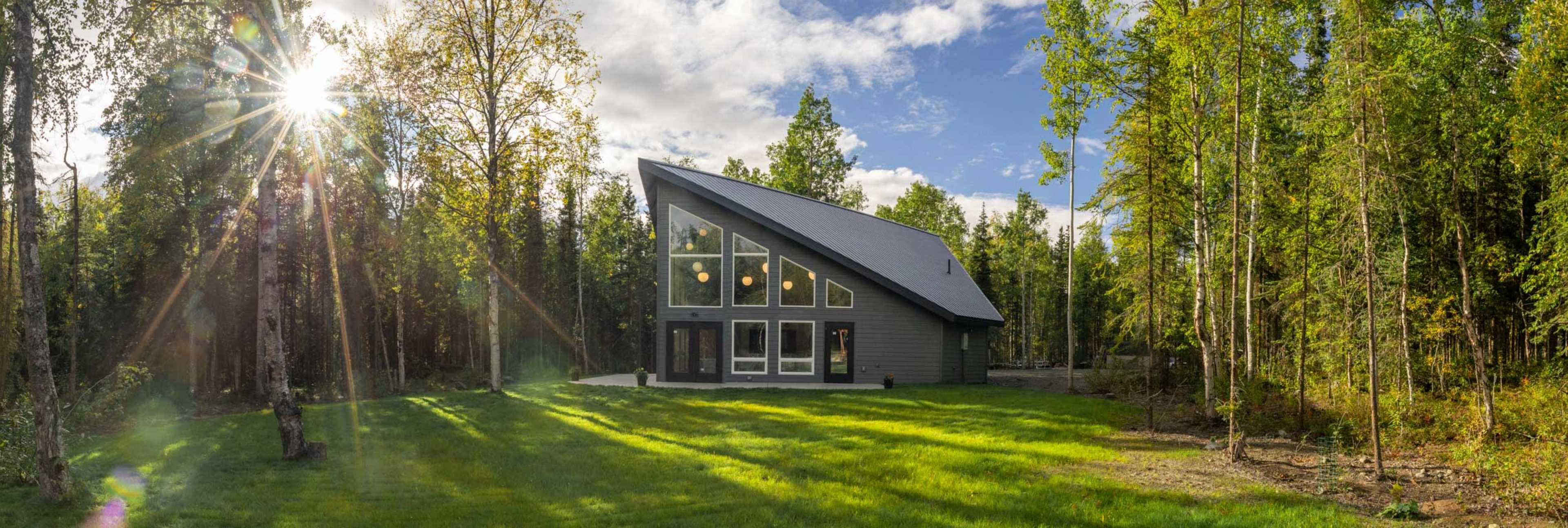 A modern, A-frame house with large windows is situated in a forest clearing, surrounded by greenery and sunlight filtering through the trees.