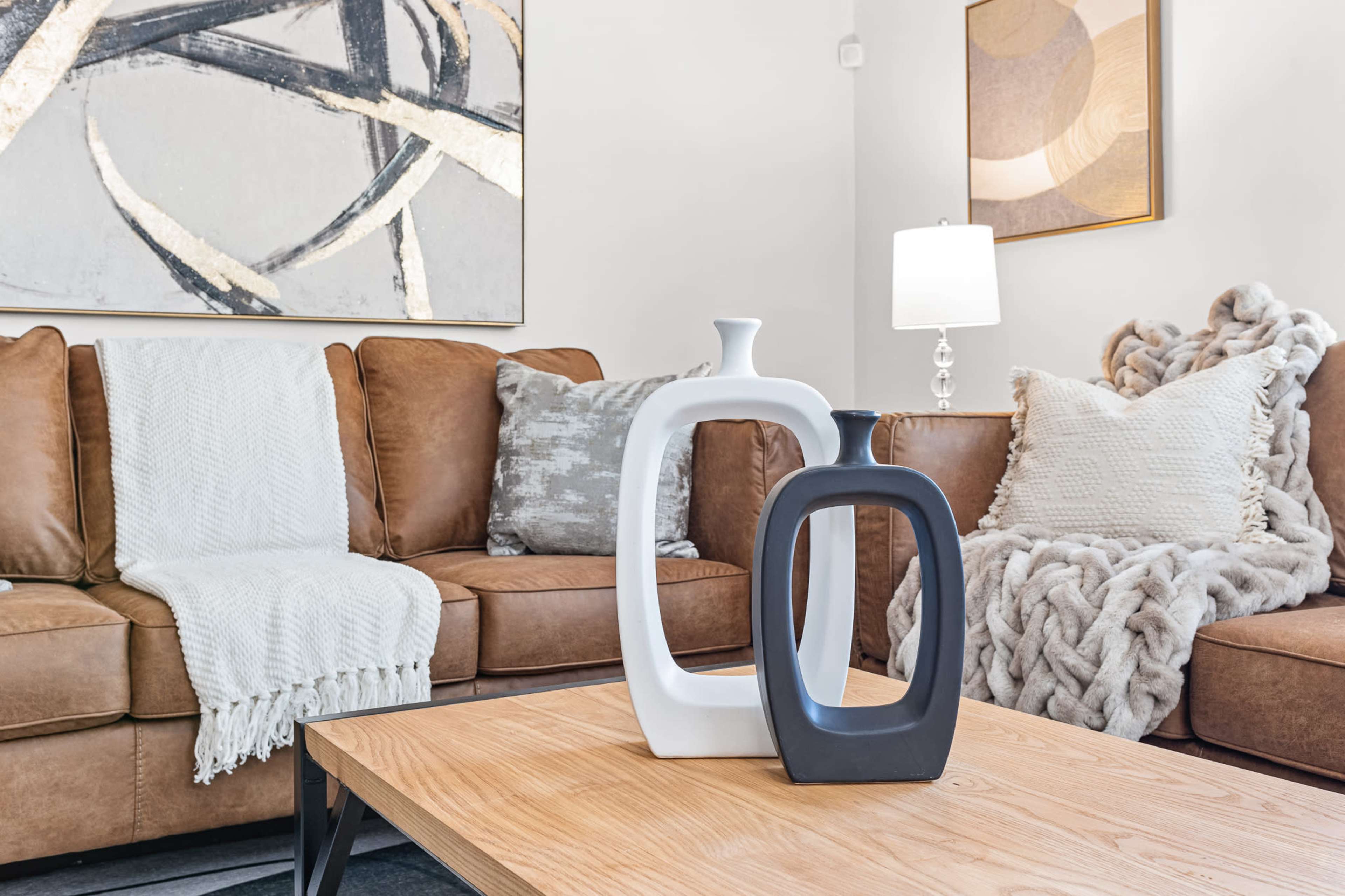 A modern living room features a brown sectional sofa, a wooden coffee table with decorative vases, and a lamp beside a textured wall art piece.