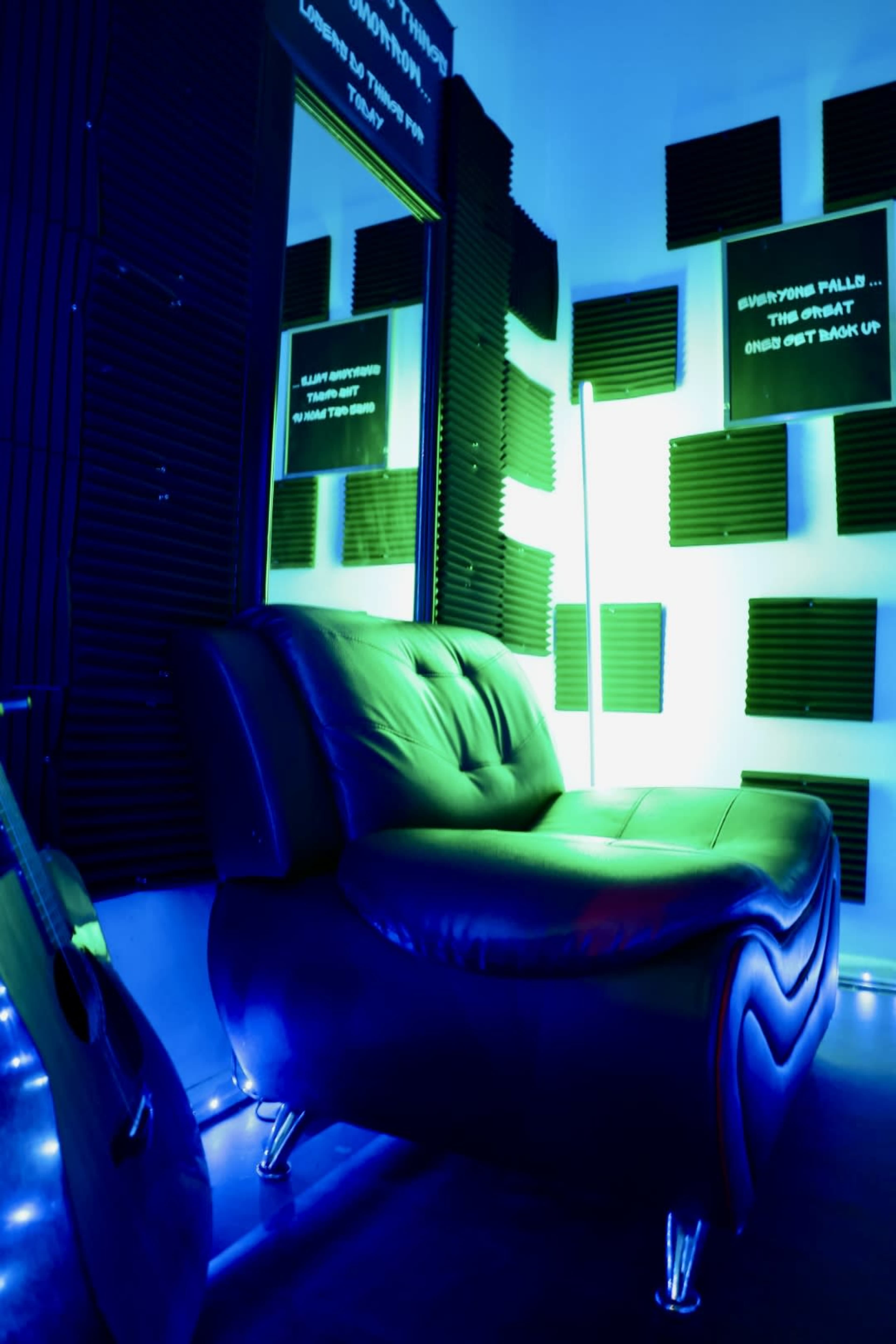 A black leather chair sits against a wall covered in soundproof panels, with green and blue lighting illuminating the space.