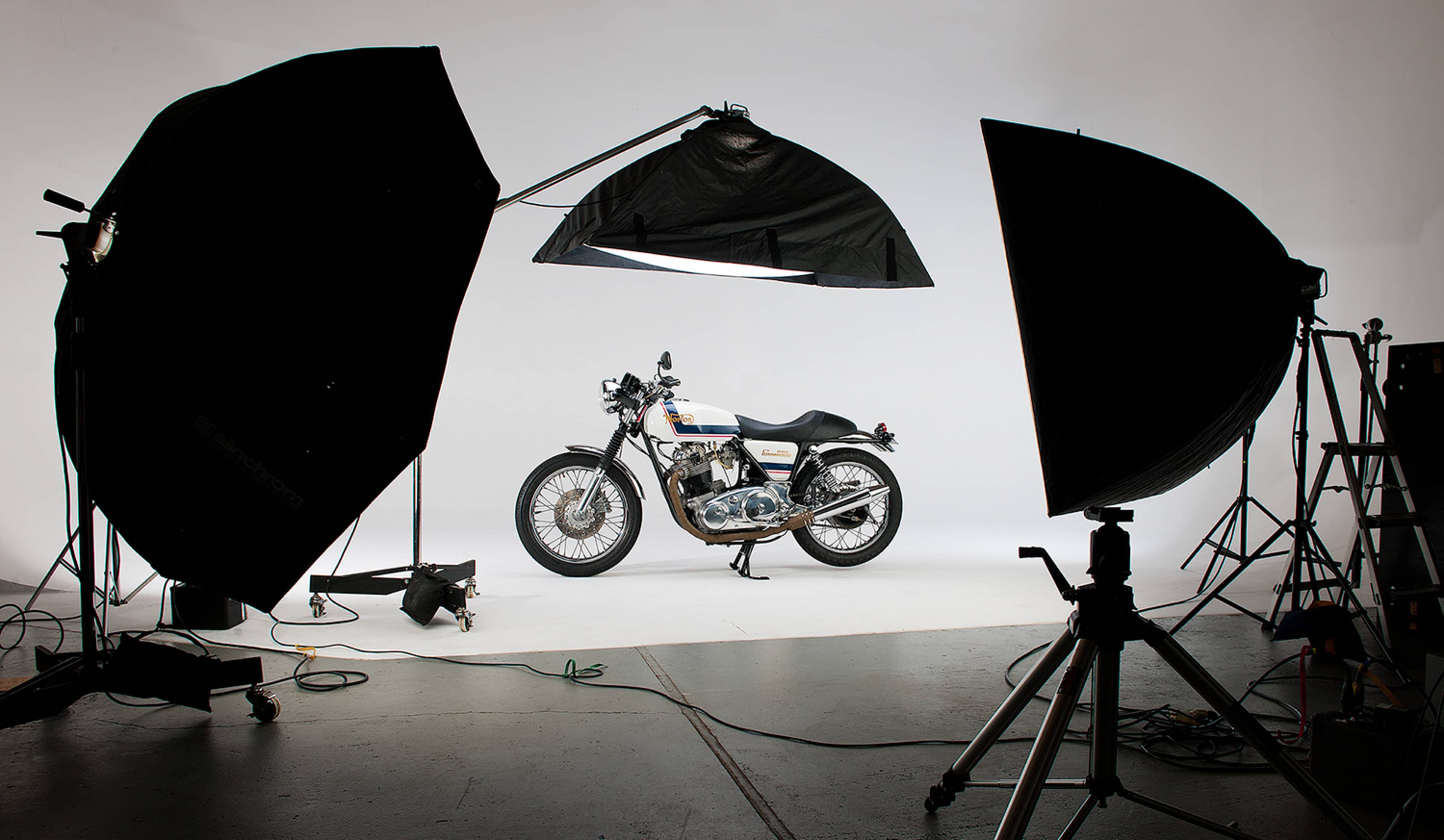 A motorcycle is positioned on a white backdrop, flanked by large light modifiers and photography equipment.