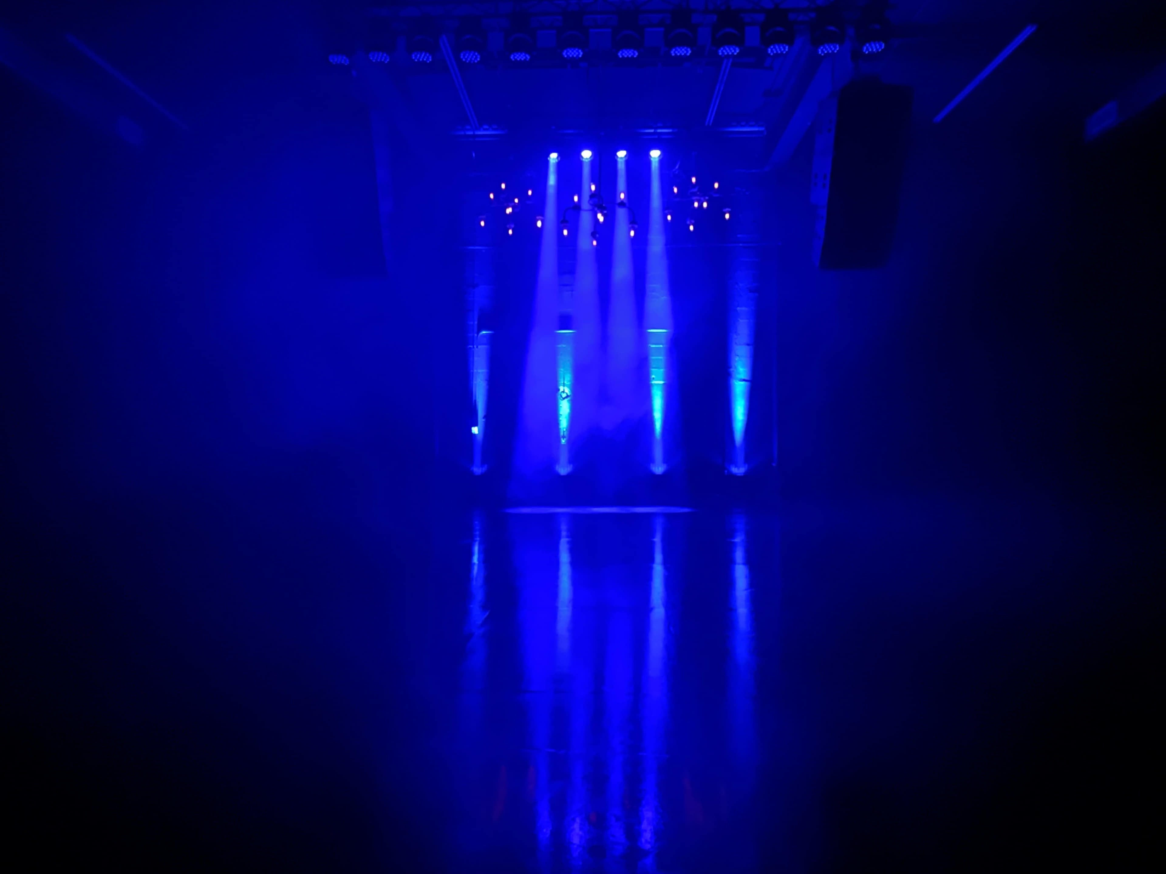 A stage is illuminated by blue lights, reflecting off a polished floor in a dim environment filled with haze.