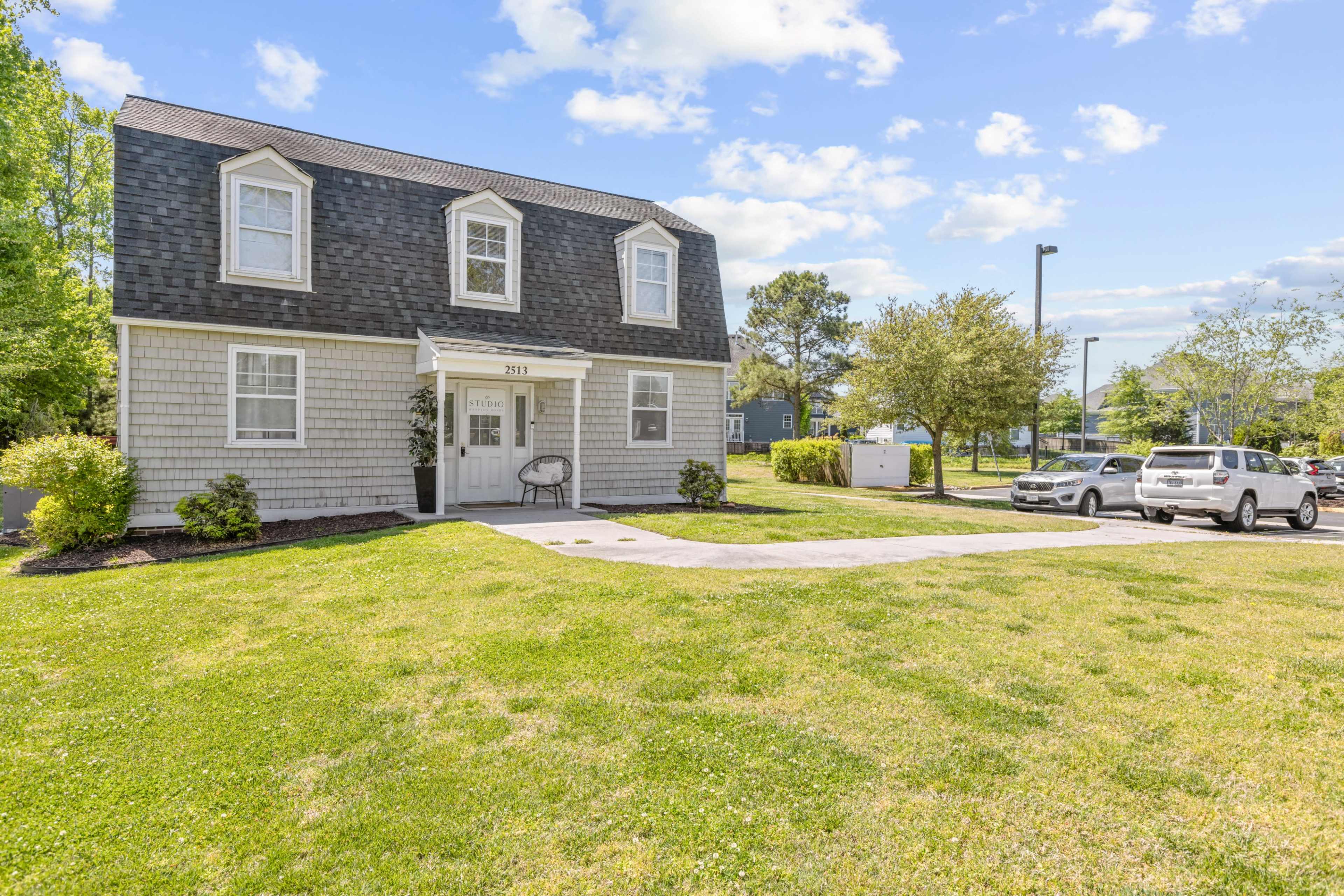 The image shows a two-story house with a shingle exterior and a spacious lawn, surrounded by green trees and parked cars.