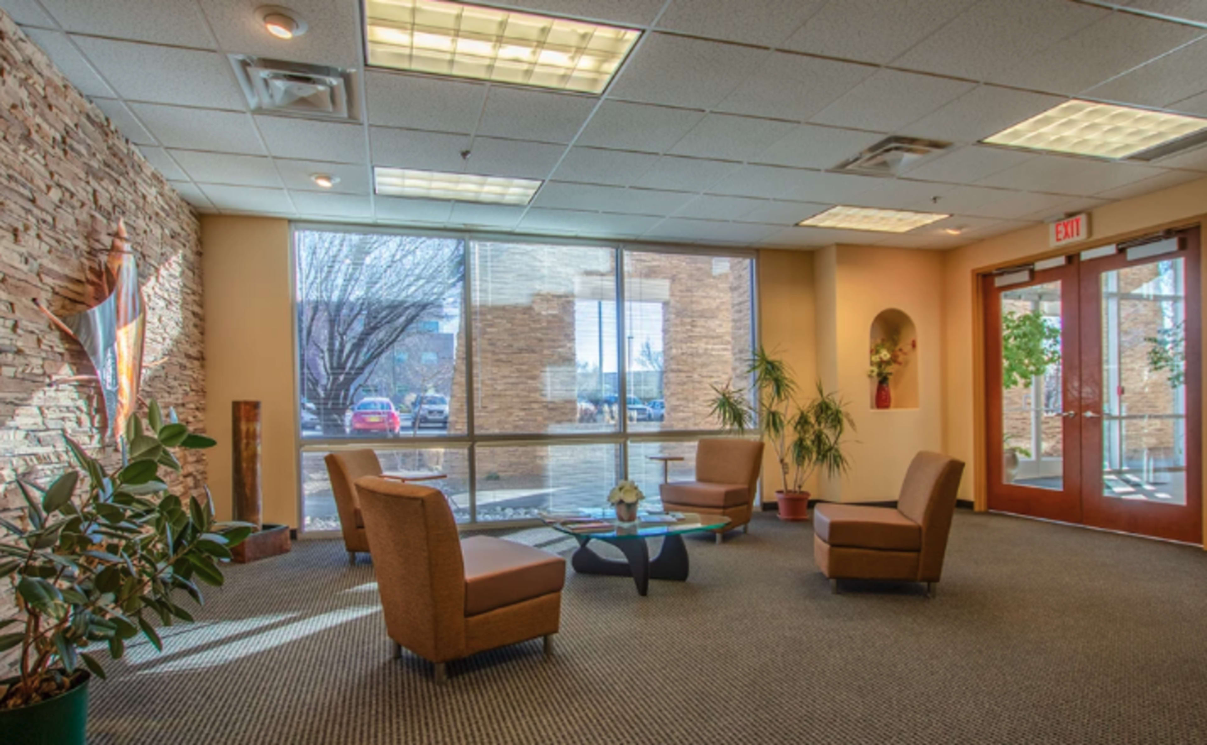 A waiting area featuring two chairs, a coffee table, and large windows with a view of an outside parking area and greenery.