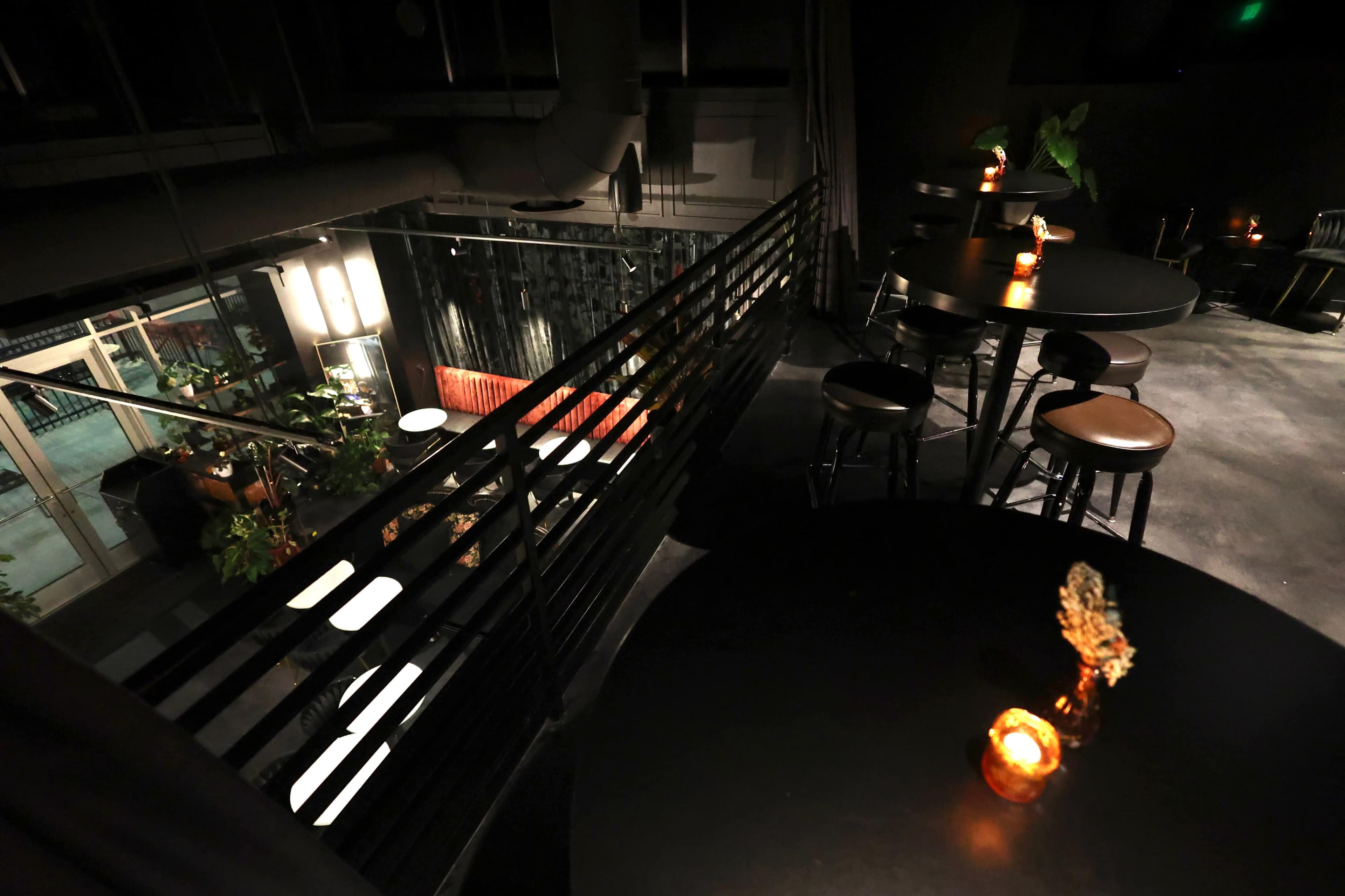 The image shows a dimly lit bar area with tables and candles, overlooking a lower seating section filled with plants and sofas.