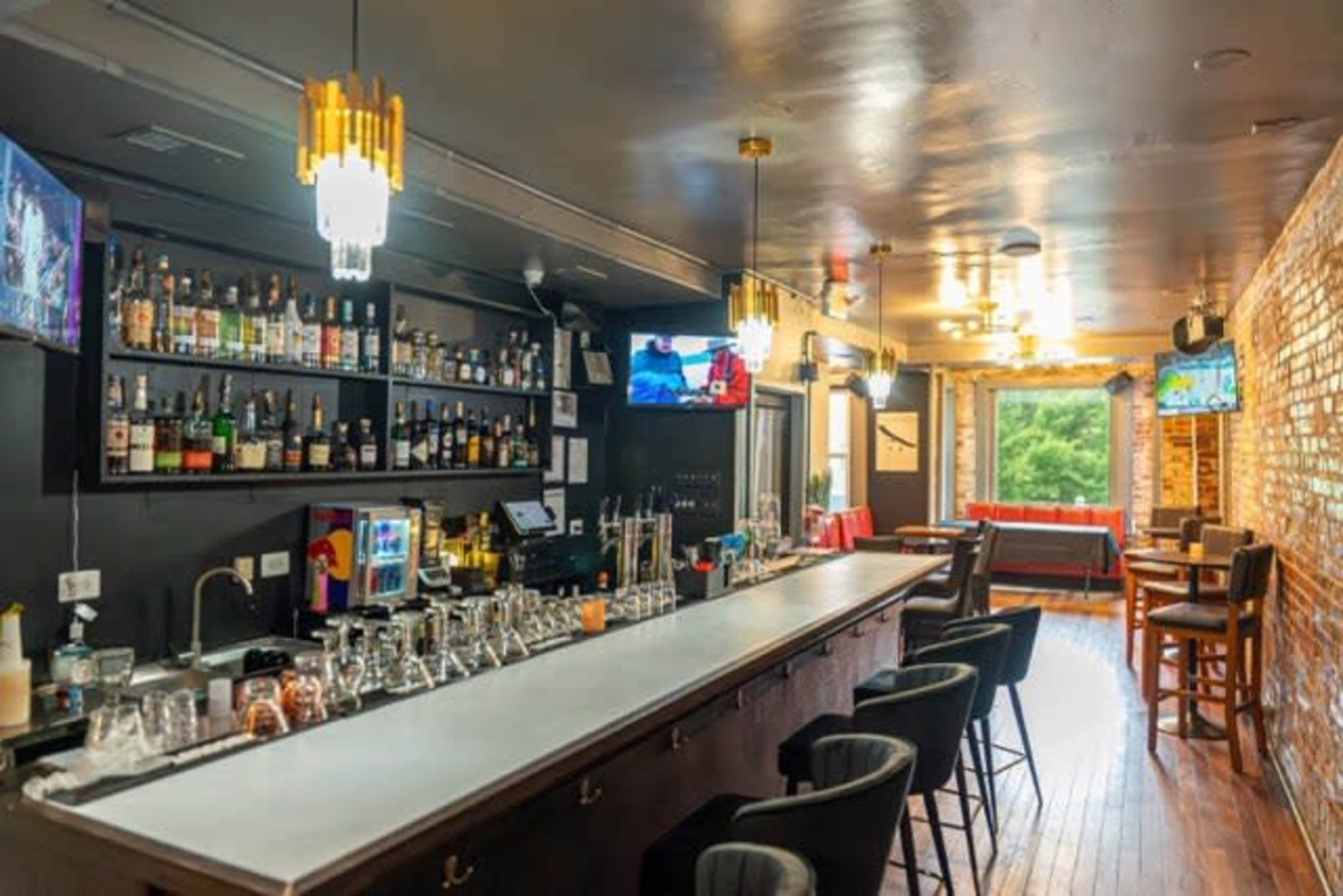 The image shows a modern bar interior with a light-colored countertop, a variety of bottles on shelves, and several bar stools lined up along the counter.