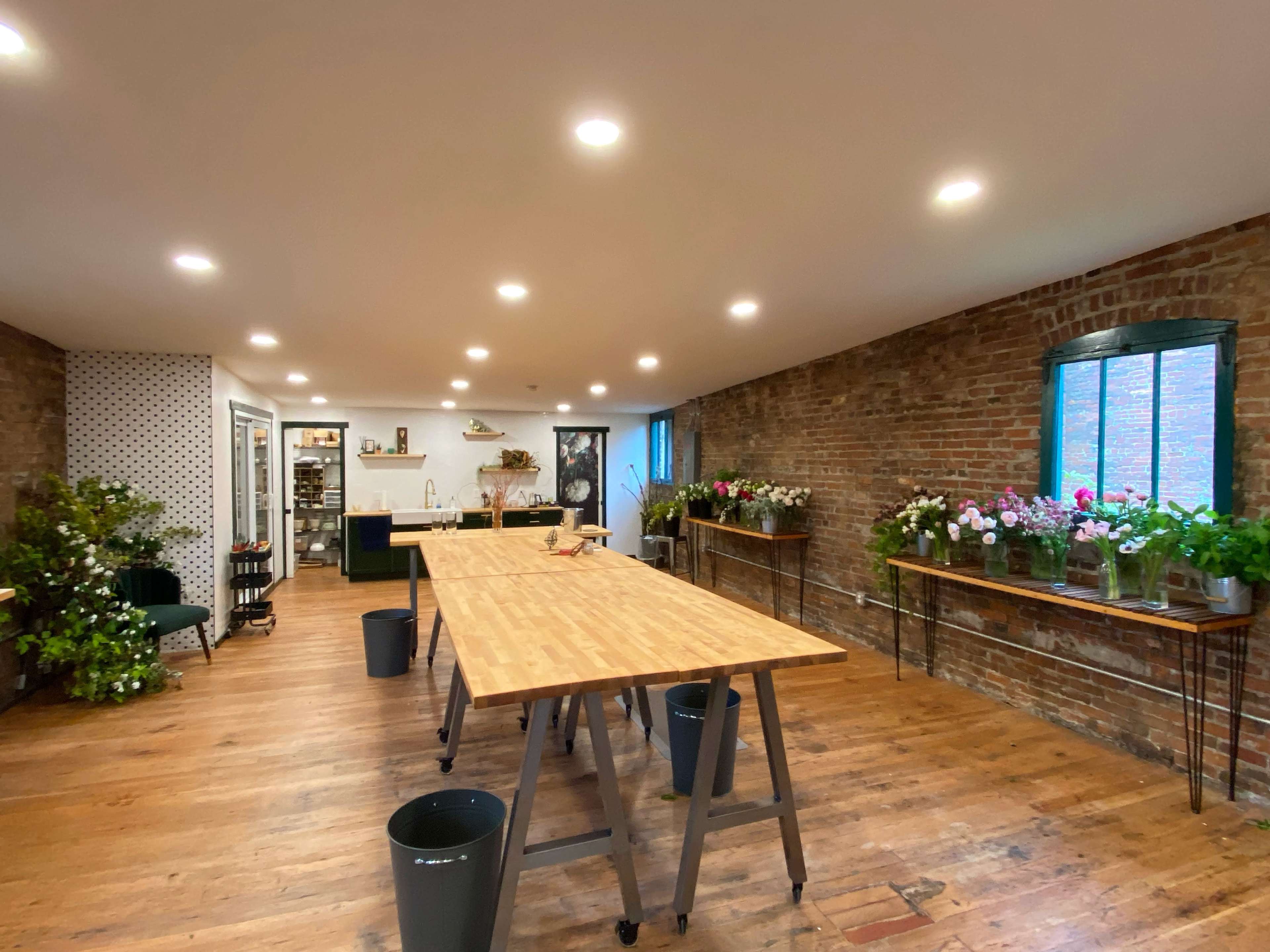 A spacious floral workshop featuring wooden tables, exposed brick walls, and various flower arrangements near the windows.