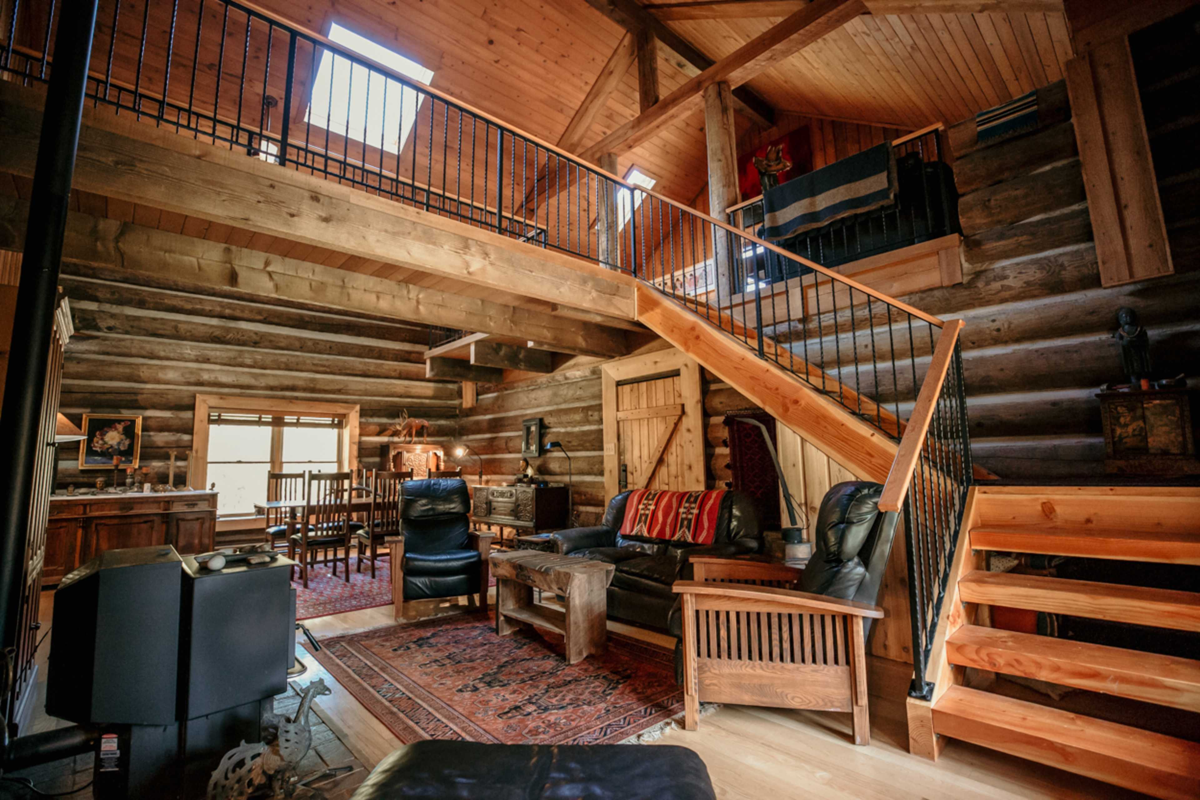 A rustic log cabin interior with a wooden staircase, leather seating, and a dining area visible in the background.