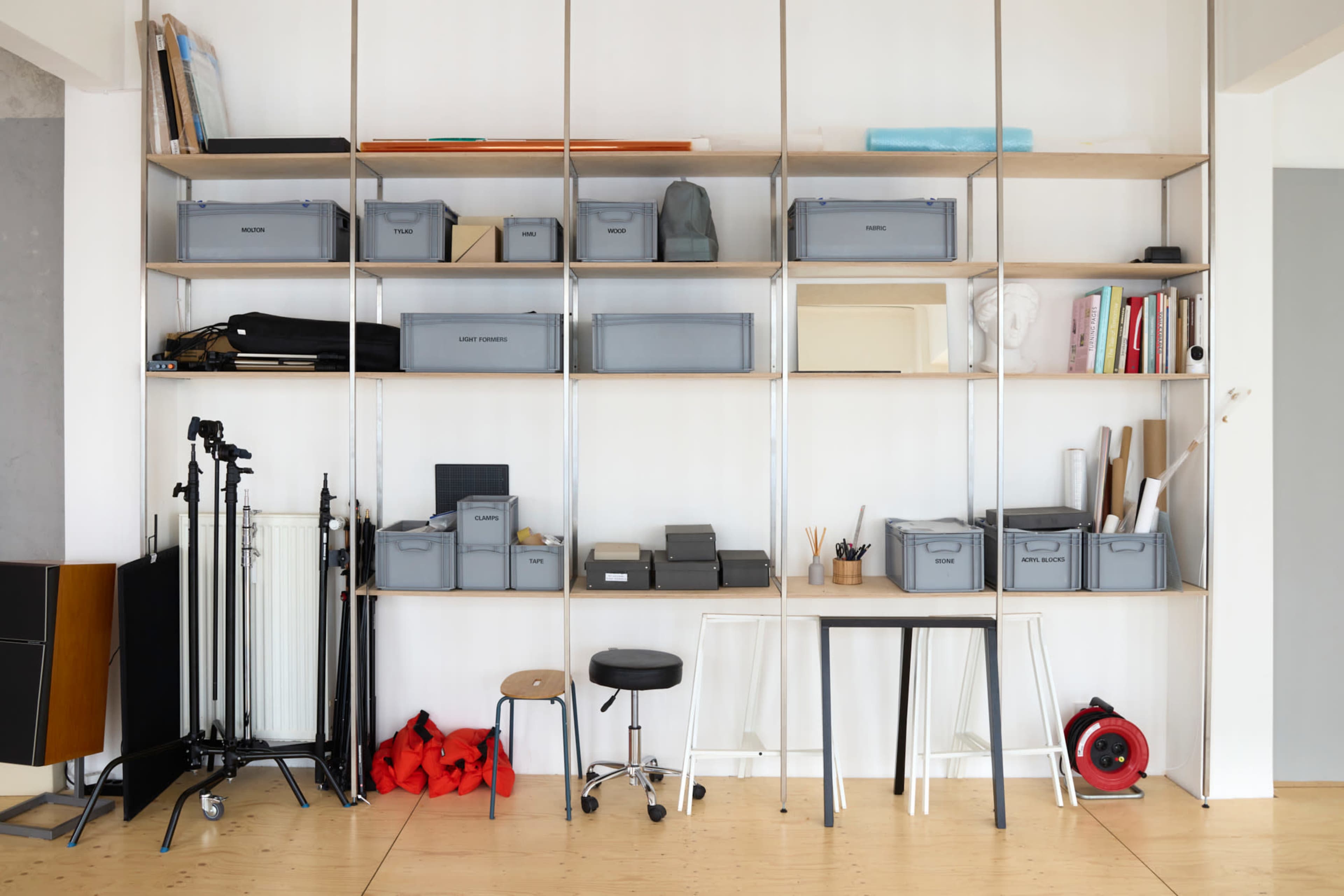 The image shows a neatly organized shelving unit filled with various storage boxes, books, and tools in a minimalist workspace.