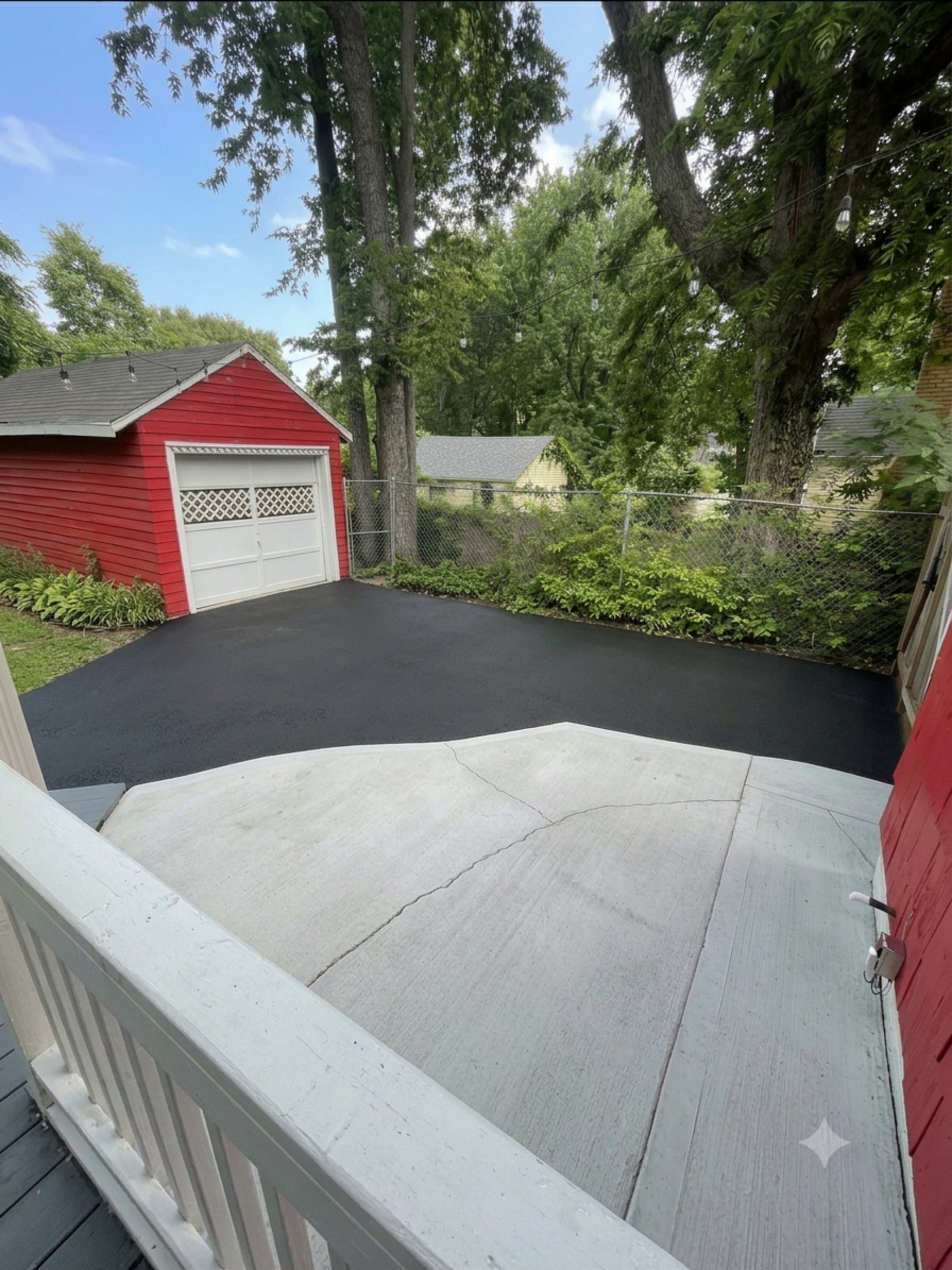 The image shows a view from a porch, featuring a freshly paved black driveway alongside a red garage and greenery in the background.