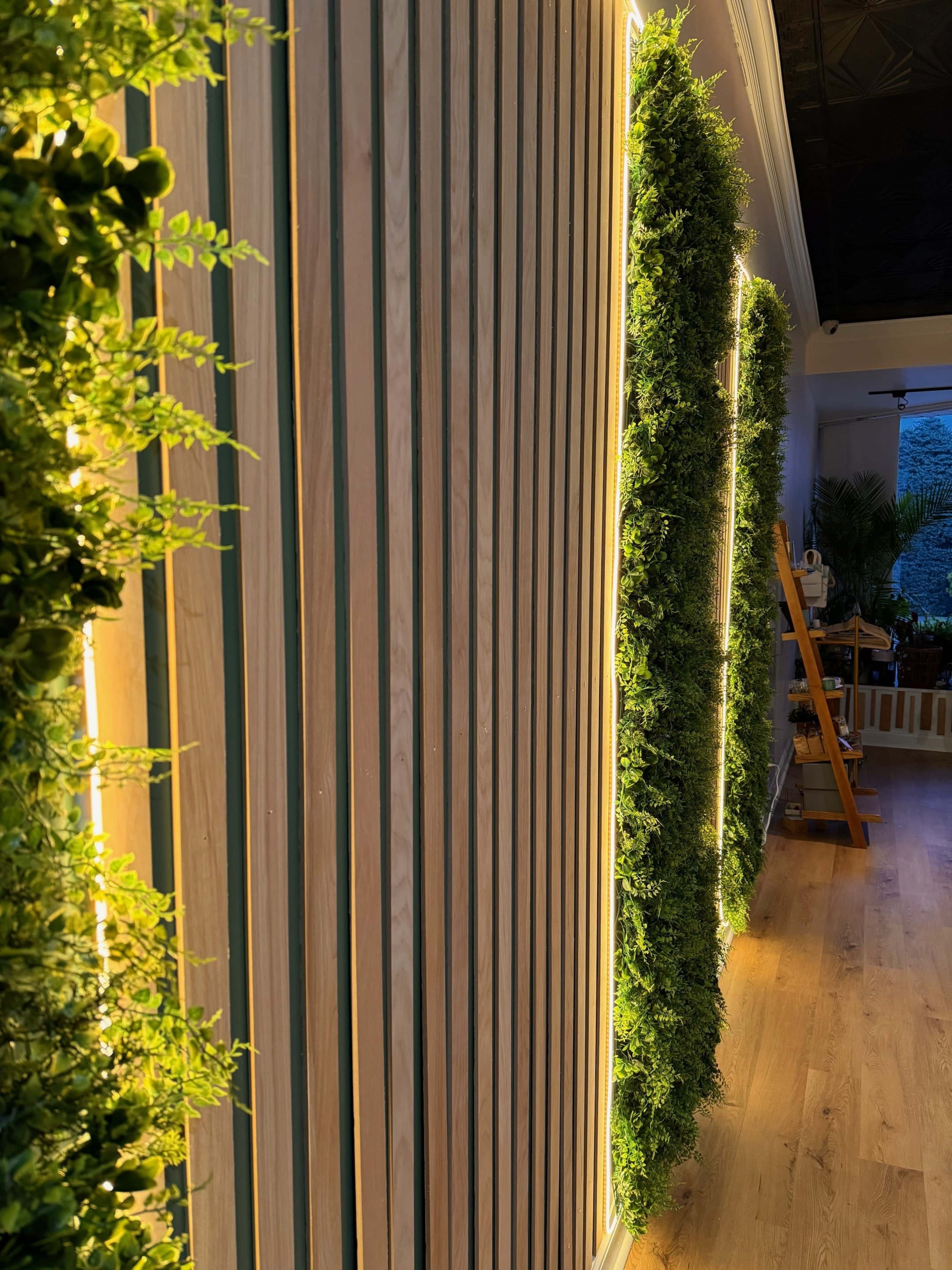 The image shows a modern interior wall featuring wooden panels flanked by vertical green plant decorations illuminated by warm lighting.