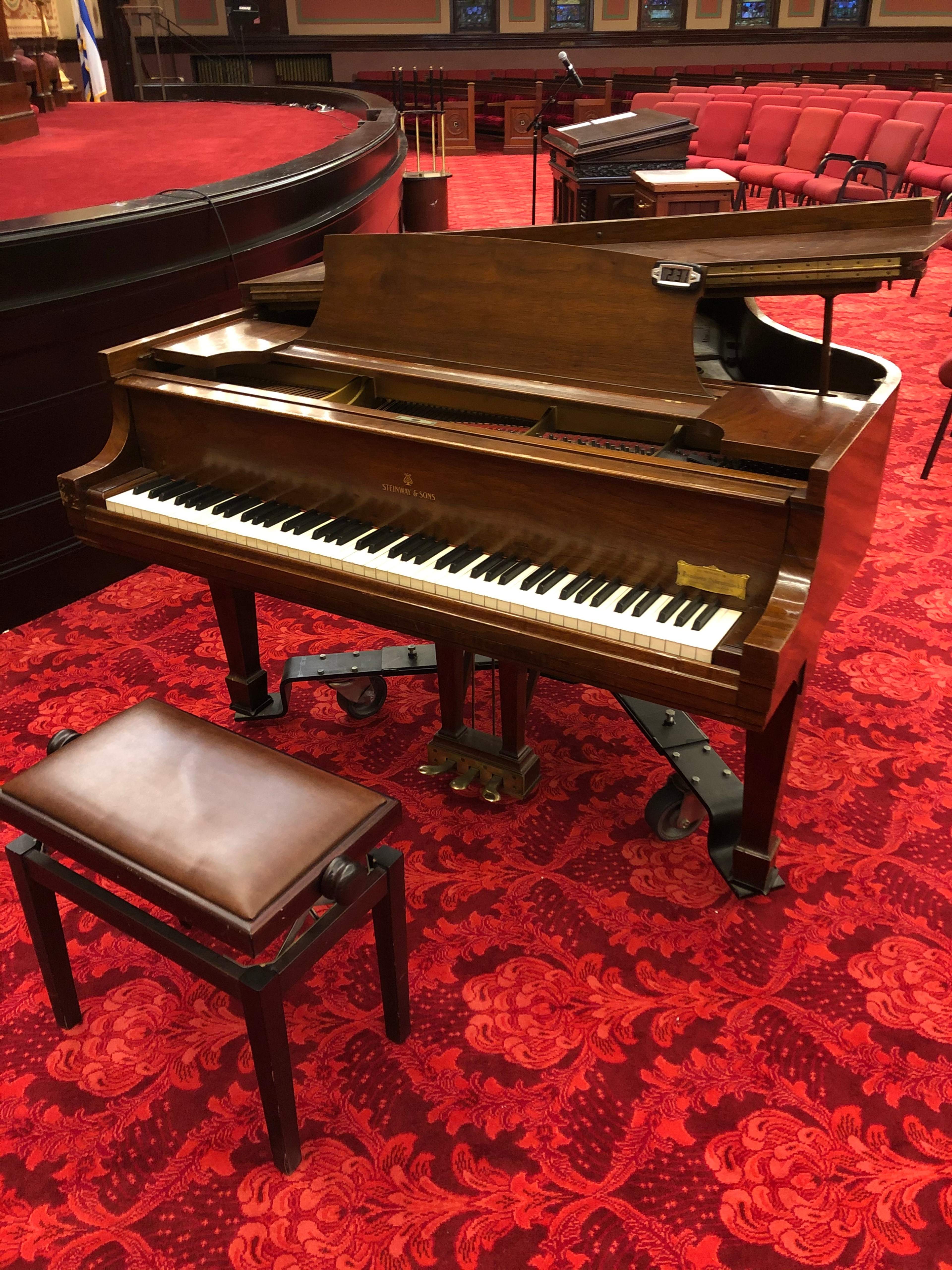 A grand piano with a matching bench is placed on a red carpet in a room with empty chairs arranged nearby.