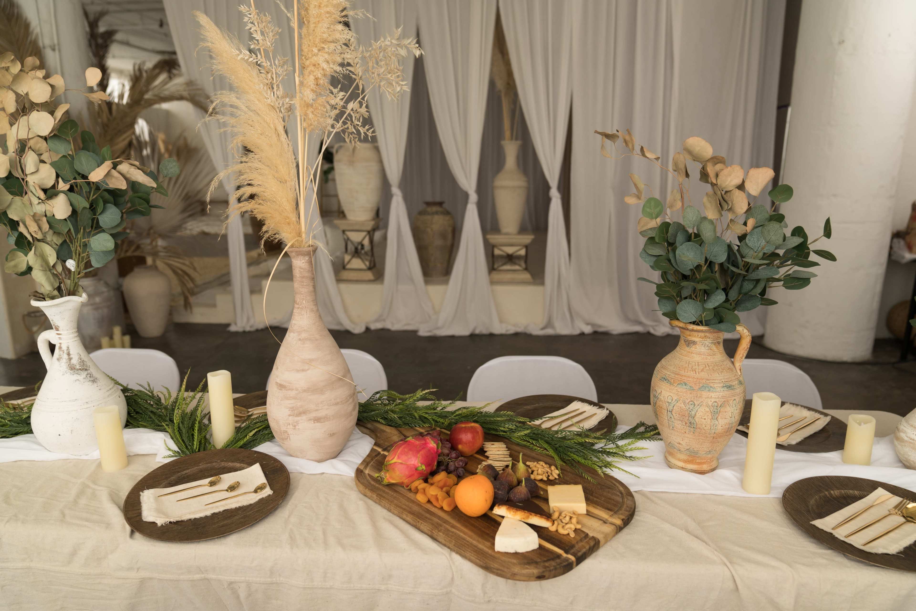 The table is set with decorative vases containing dried foliage, wooden trays featuring an assortment of fruits and cheeses, and neatly arranged tableware.