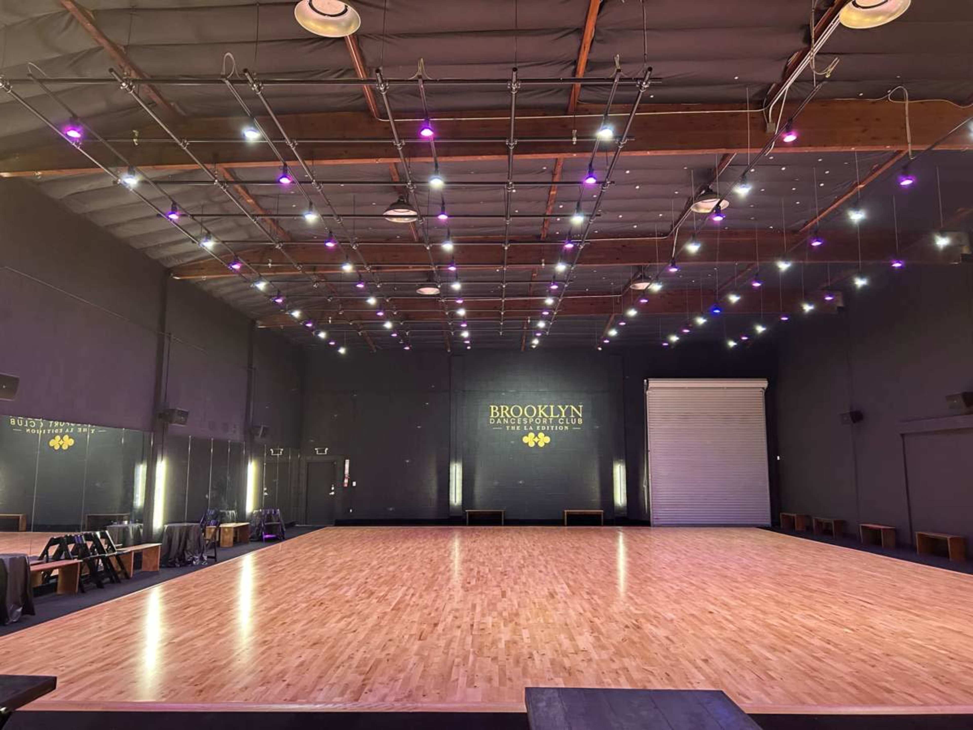 The image shows a spacious dance studio with a polished wooden floor, brightly lit by overhead lights, and a mirrored wall on one side.
