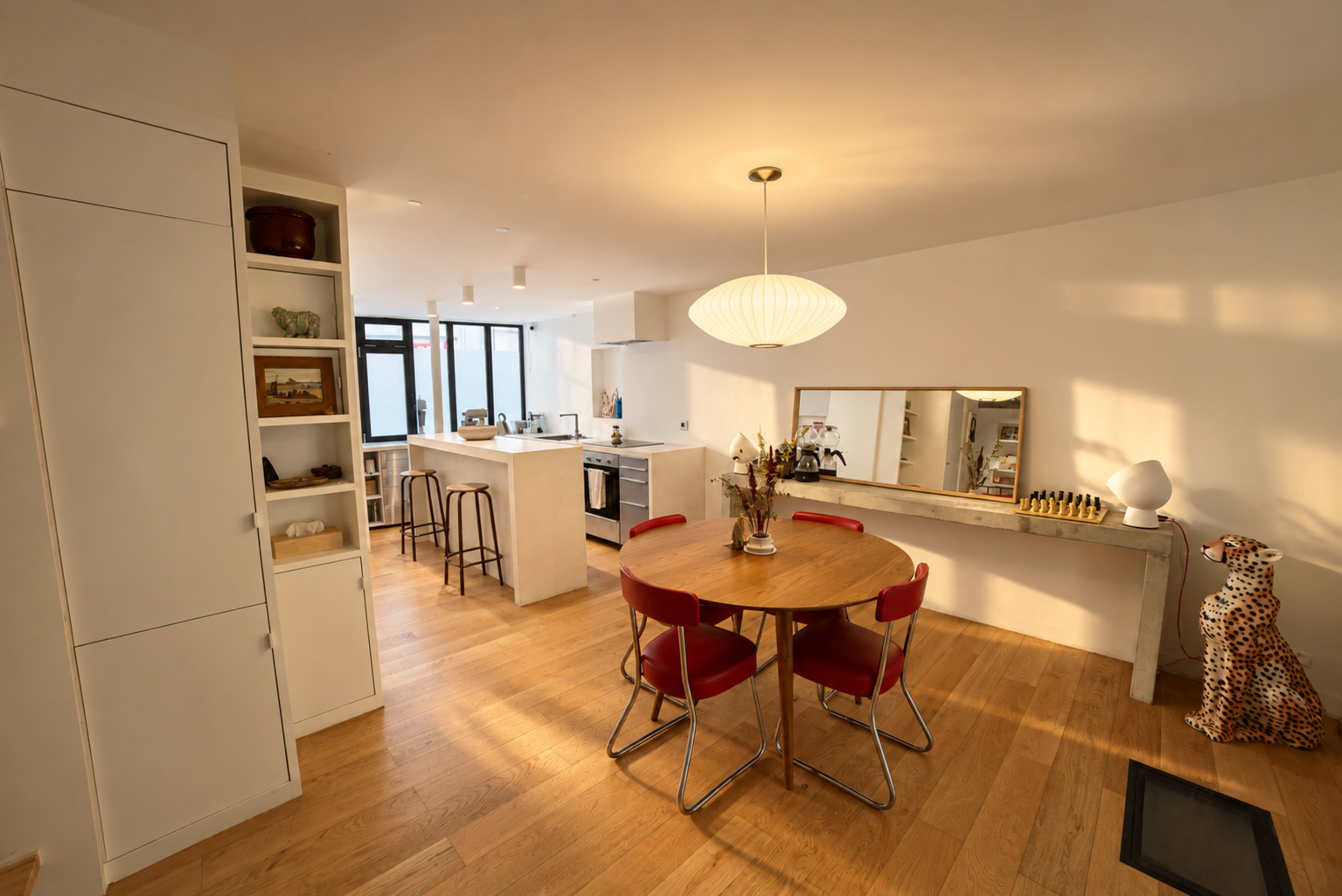 A bright dining area features a round wooden table surrounded by red chairs, with a kitchen visible in the background.