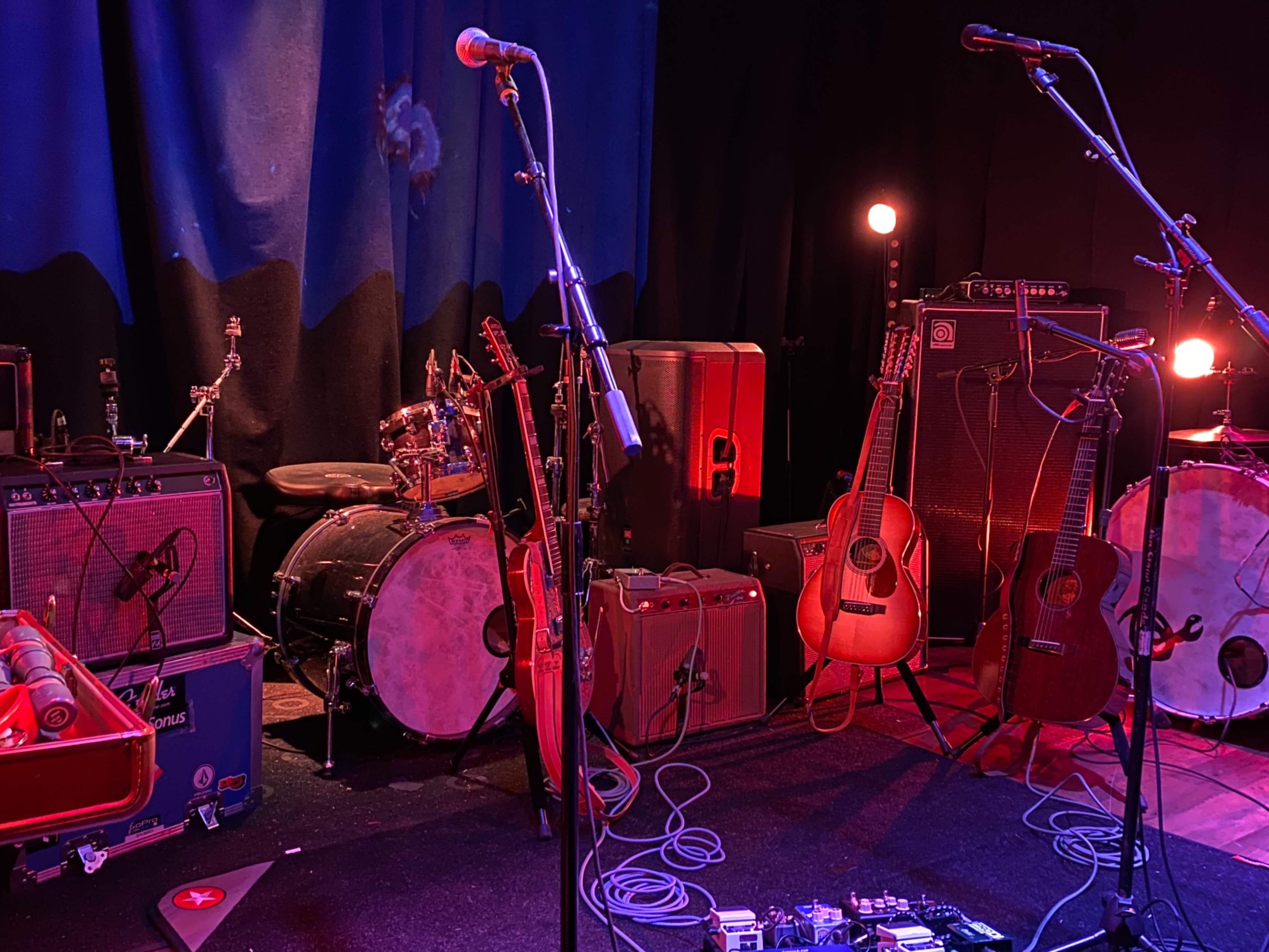 The stage is set with various musical instruments, including guitars, amplifiers, a drum kit, and microphones, ready for performance.
