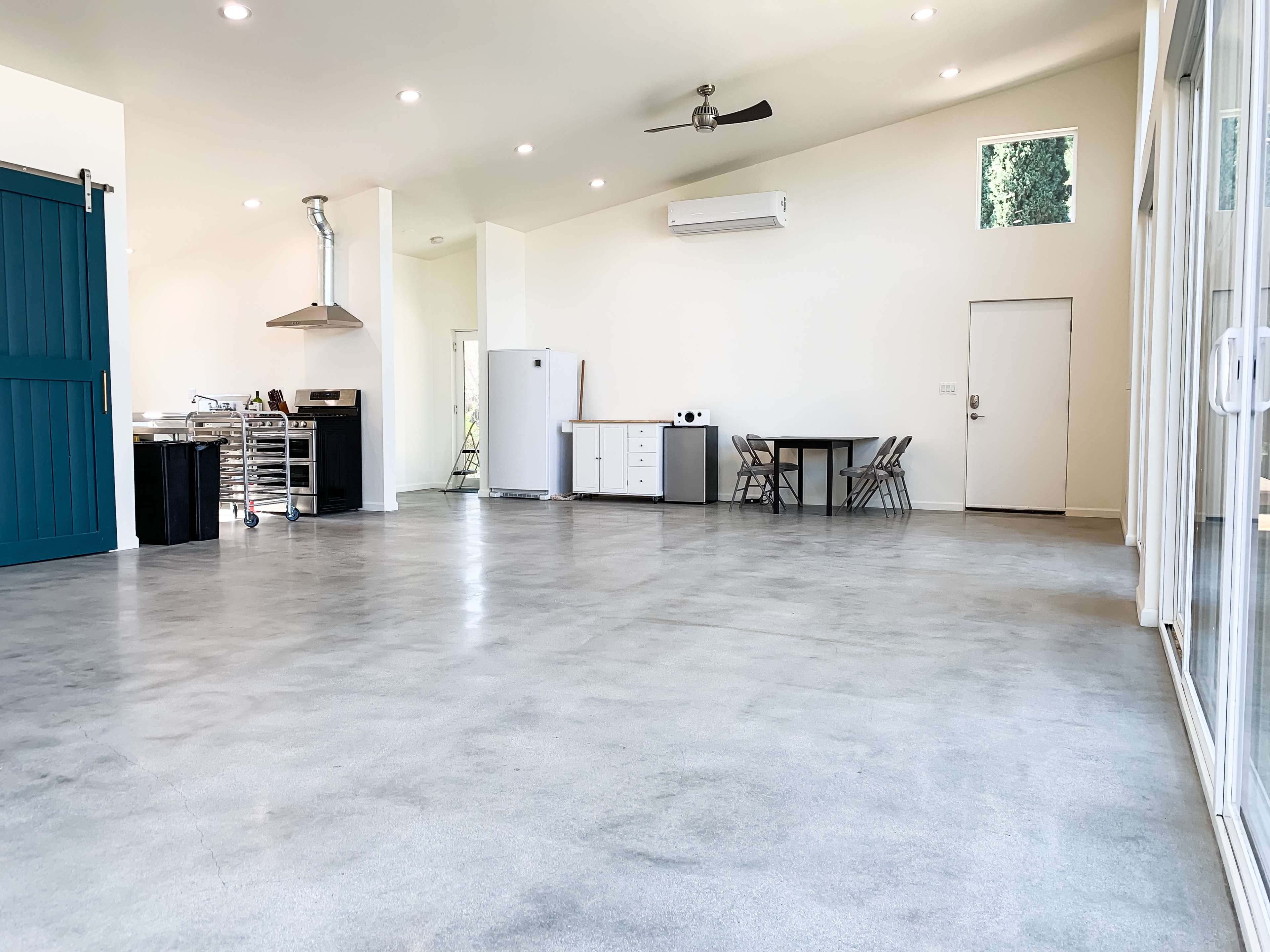 An open-concept interior with polished concrete floors, a kitchen area to the left featuring a large blue door, and a dining table set against a wall with windows on the right.